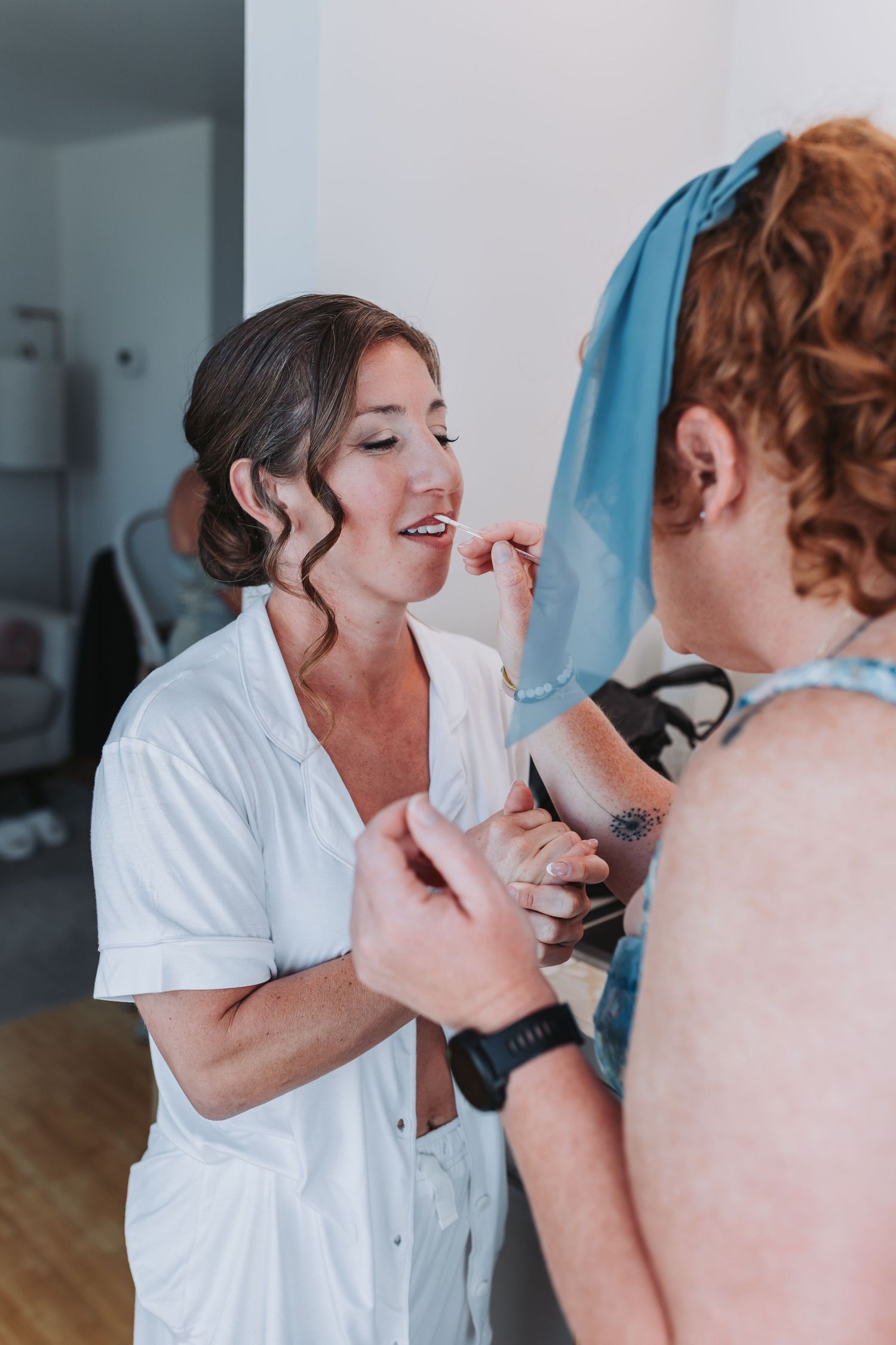 Bride in white robe smiling during wedding preparations while getting ready with a bridesmaid.
