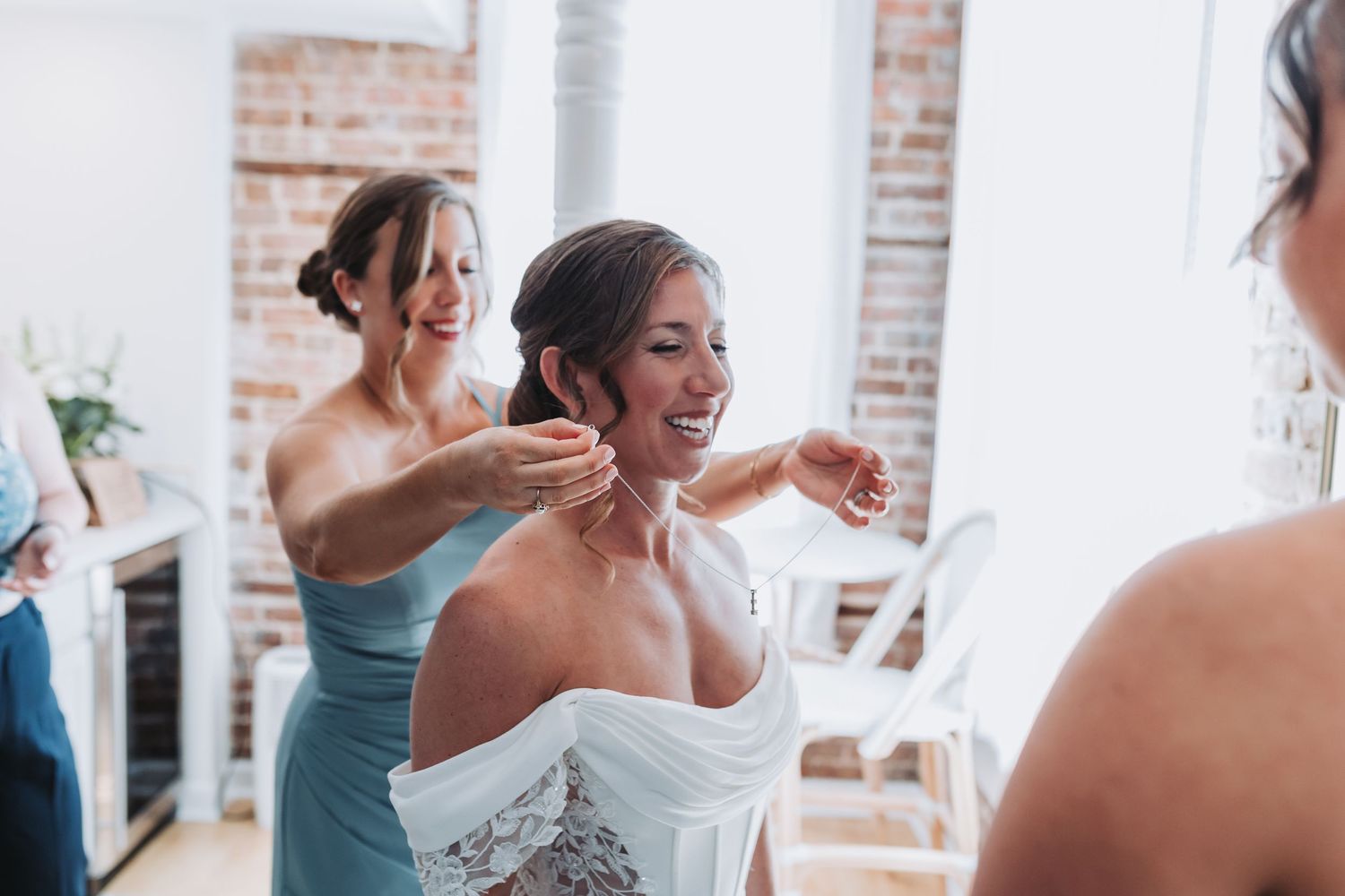 Bridesmaids helping smiling bride with her necklace during wedding day preparations indoors.