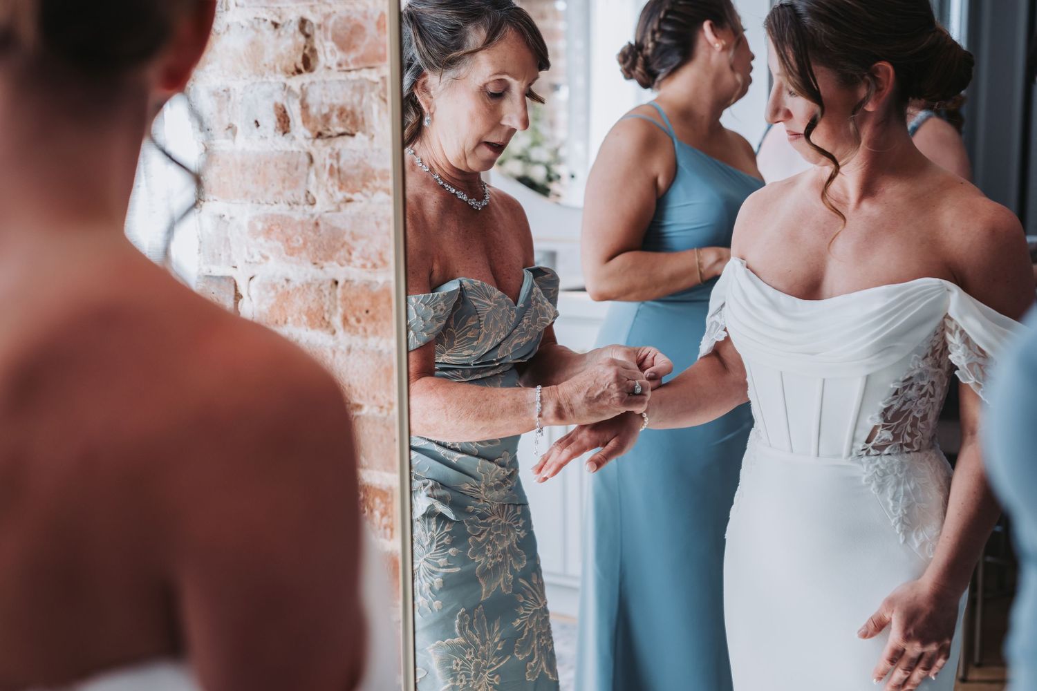 Mother helping bride with wedding dress details near mirror with bridesmaids.