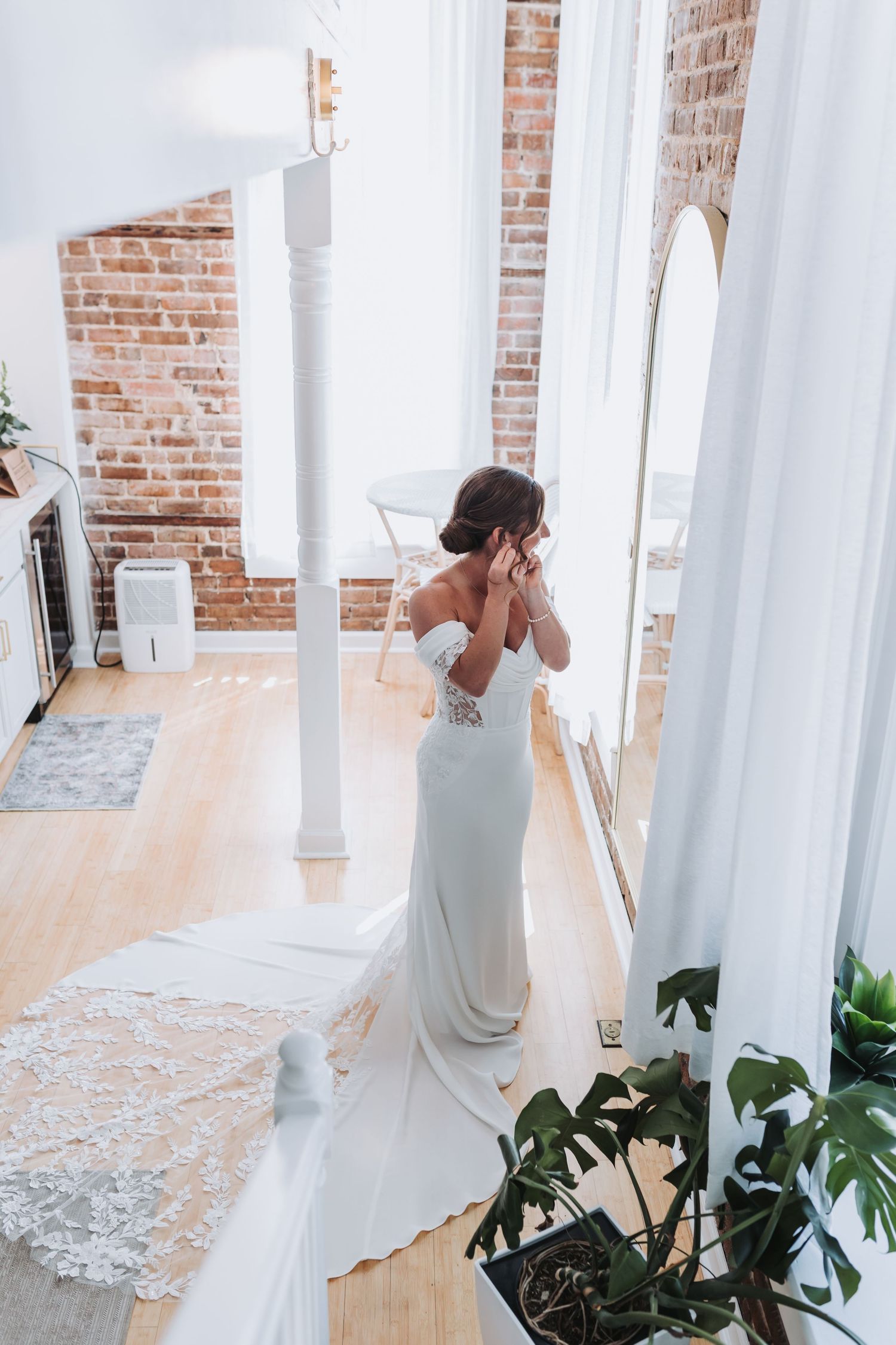 Bride in off-shoulder white gown adjusting earring near sunlit window with brick wall.
