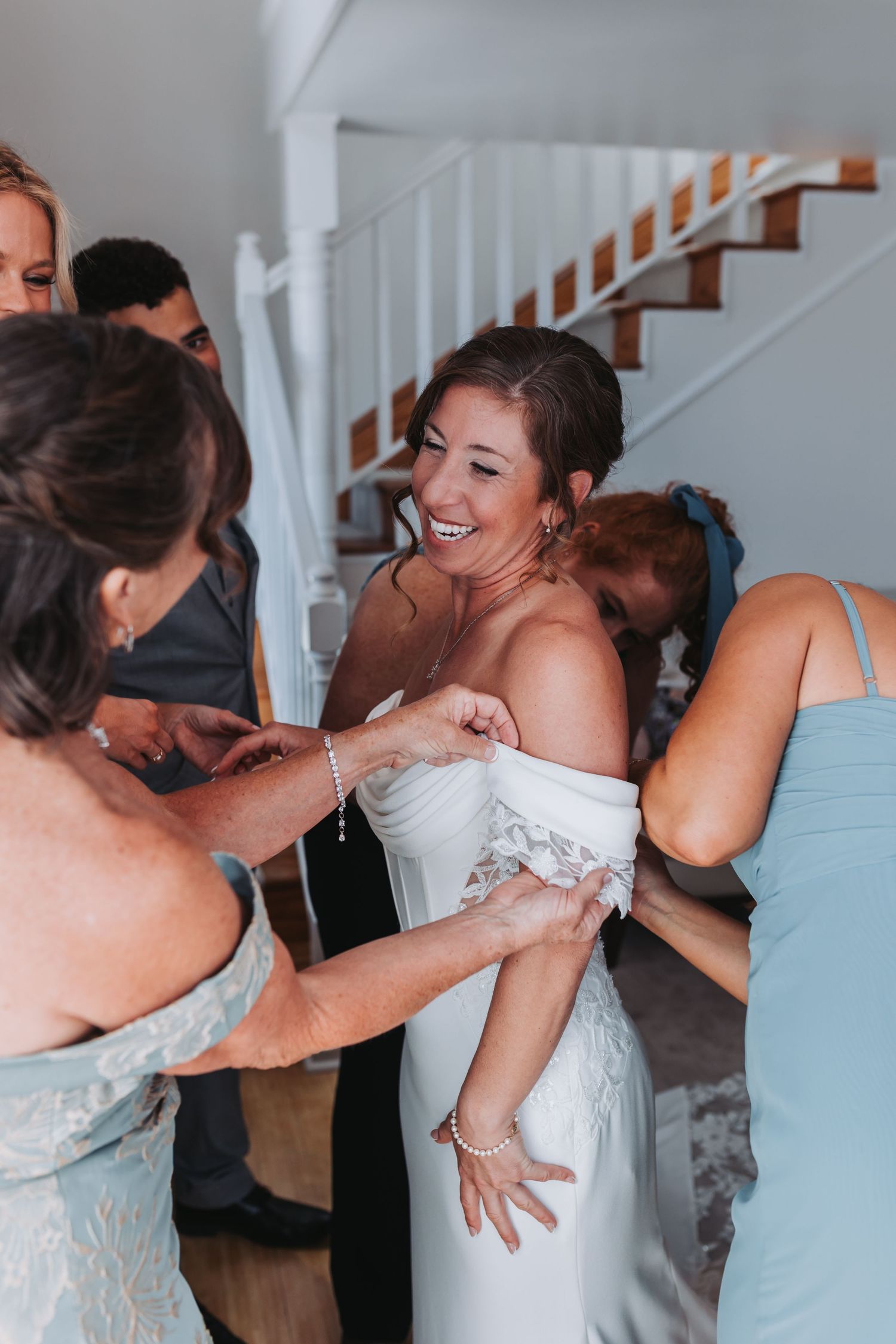 Laughing bride in off-shoulder gown gets dressed with help from bridesmaids in blue near staircase.