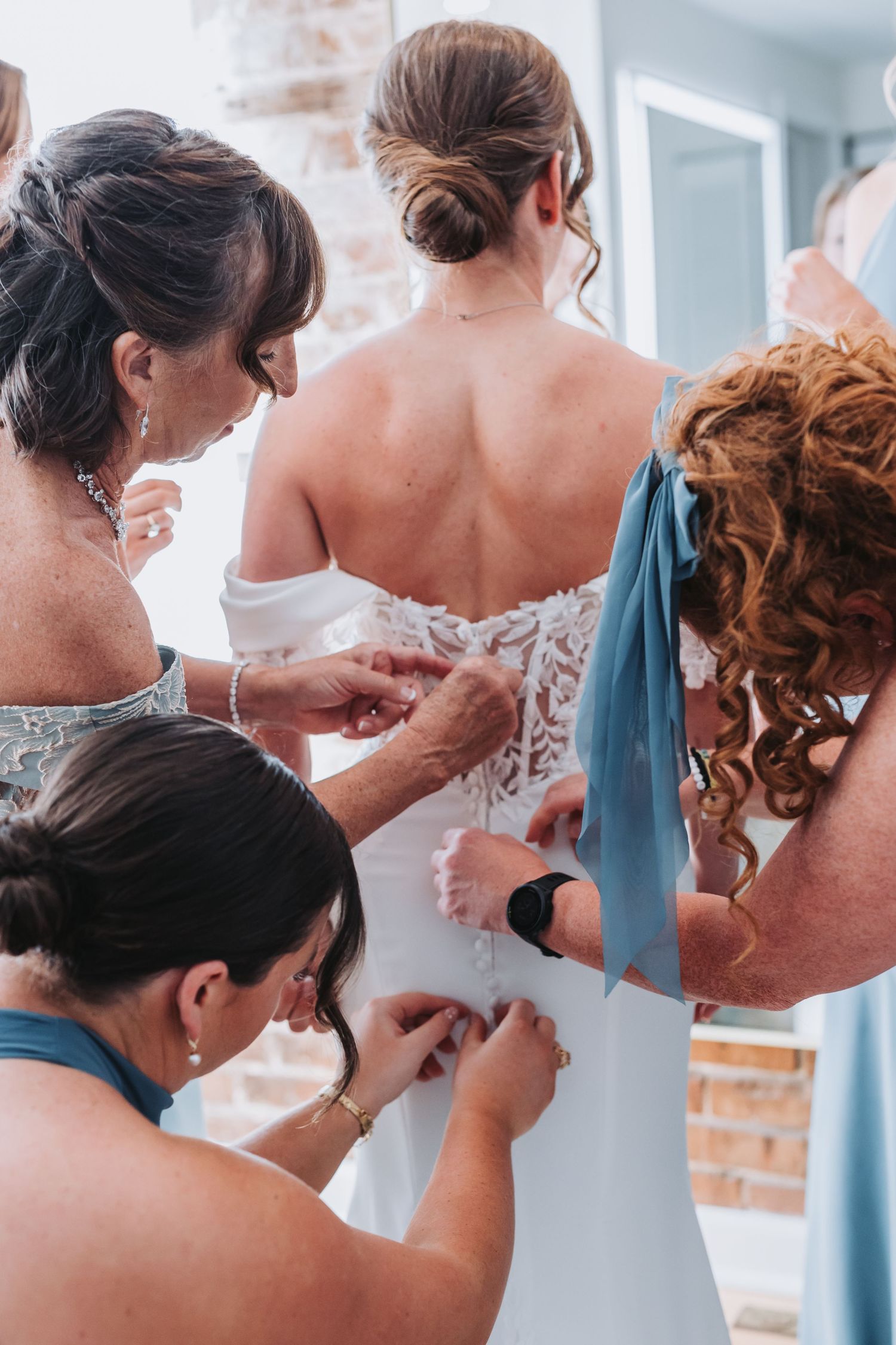 Three women carefully buttoning the back of a lace wedding dress.