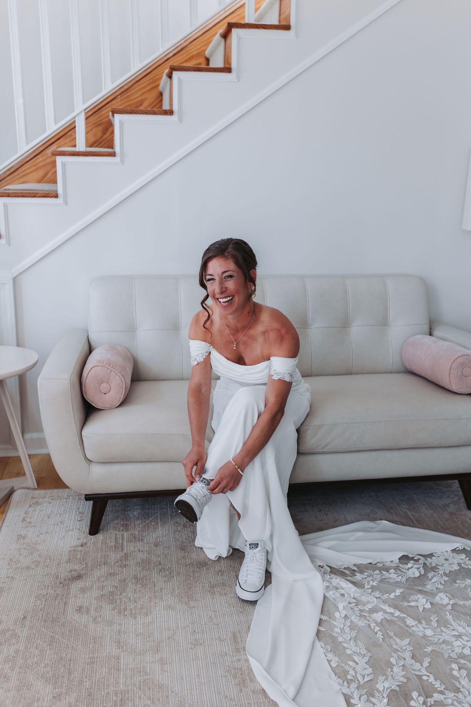 Smiling bride in off-shoulder gown putting on sneakers on white couch.
