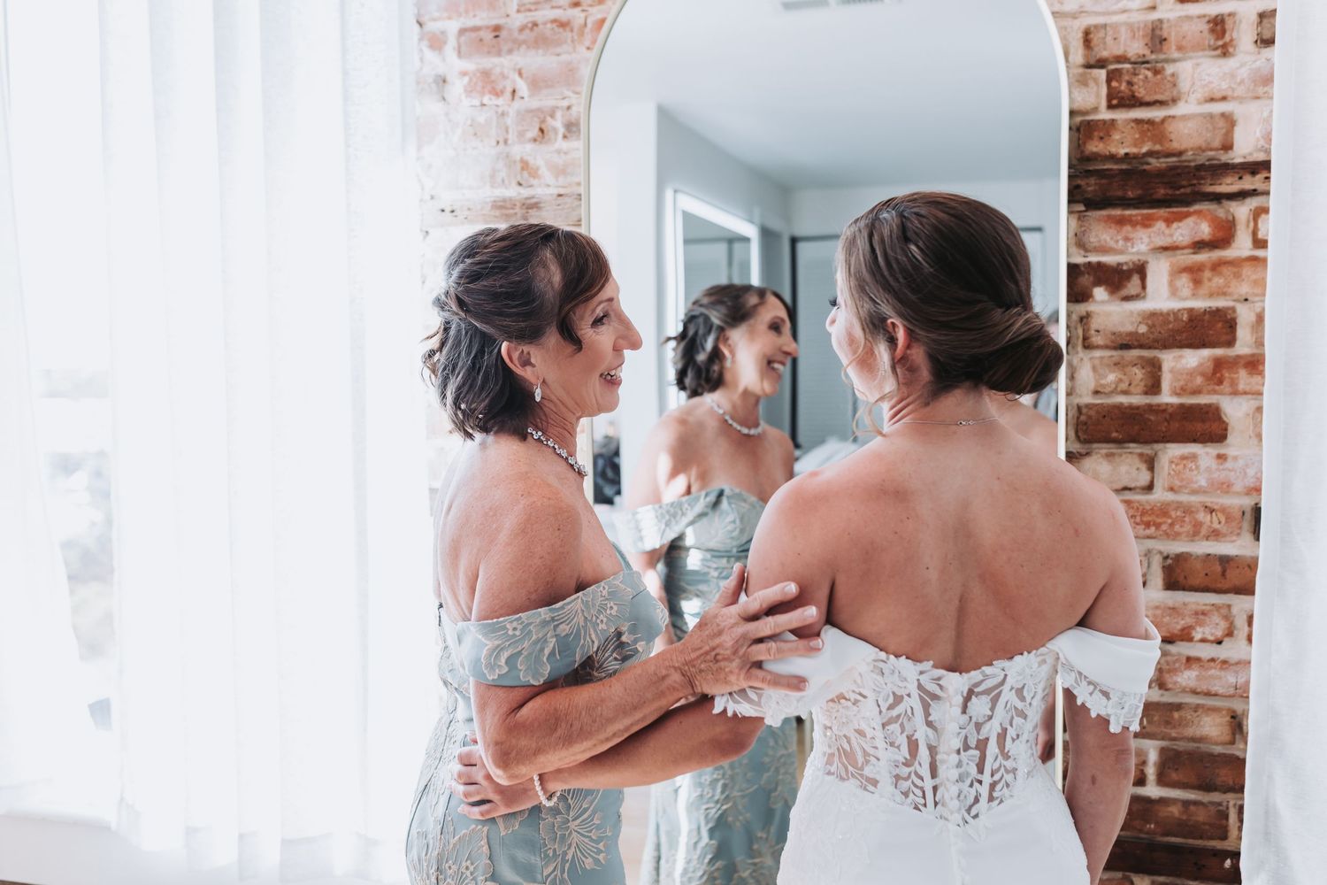 Emotional mother helping bride with lace wedding dress near mirror with brick wall backdrop.