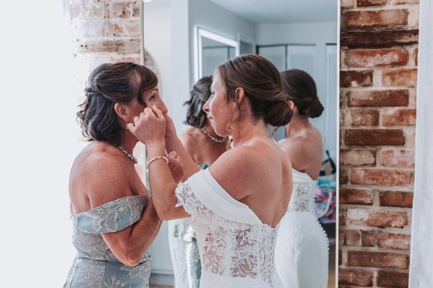 Emotional mother helping bride with earrings during wedding preparations in front of brick wall.
