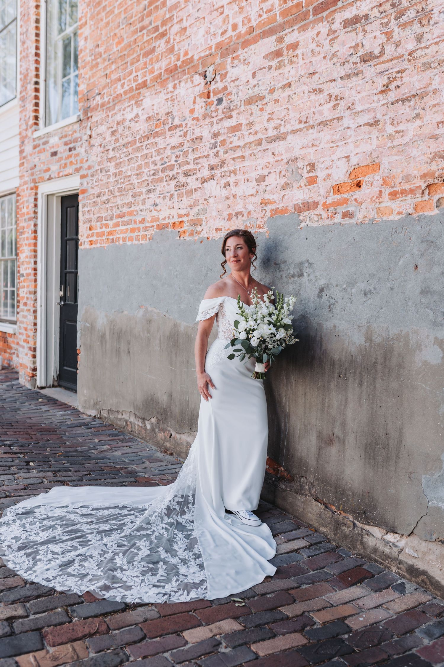 Bride in off-shoulder white gown with lace train holding bouquet against brick wall.