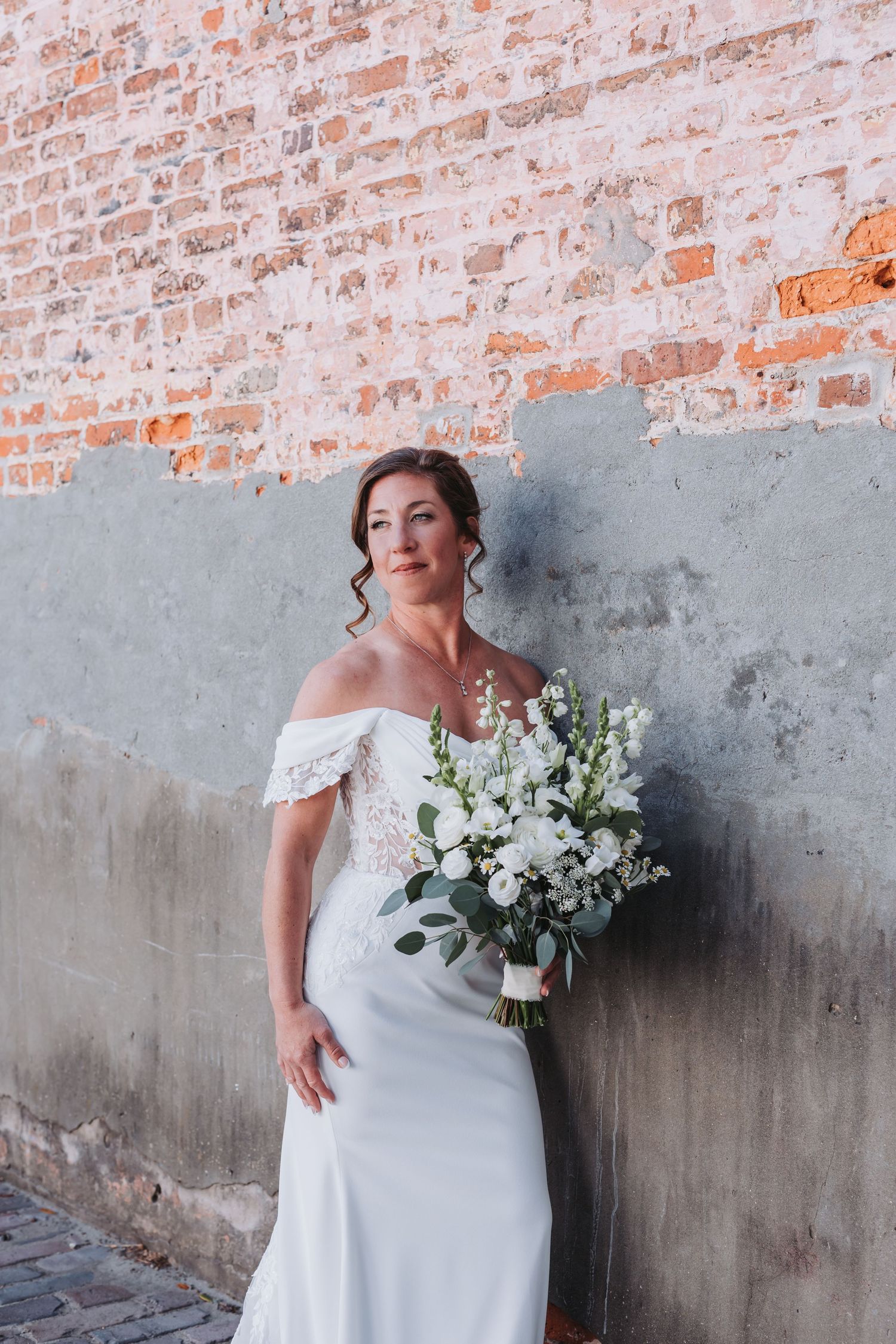 Bride in white off-shoulder gown holding white floral bouquet against rustic brick and concrete wall.