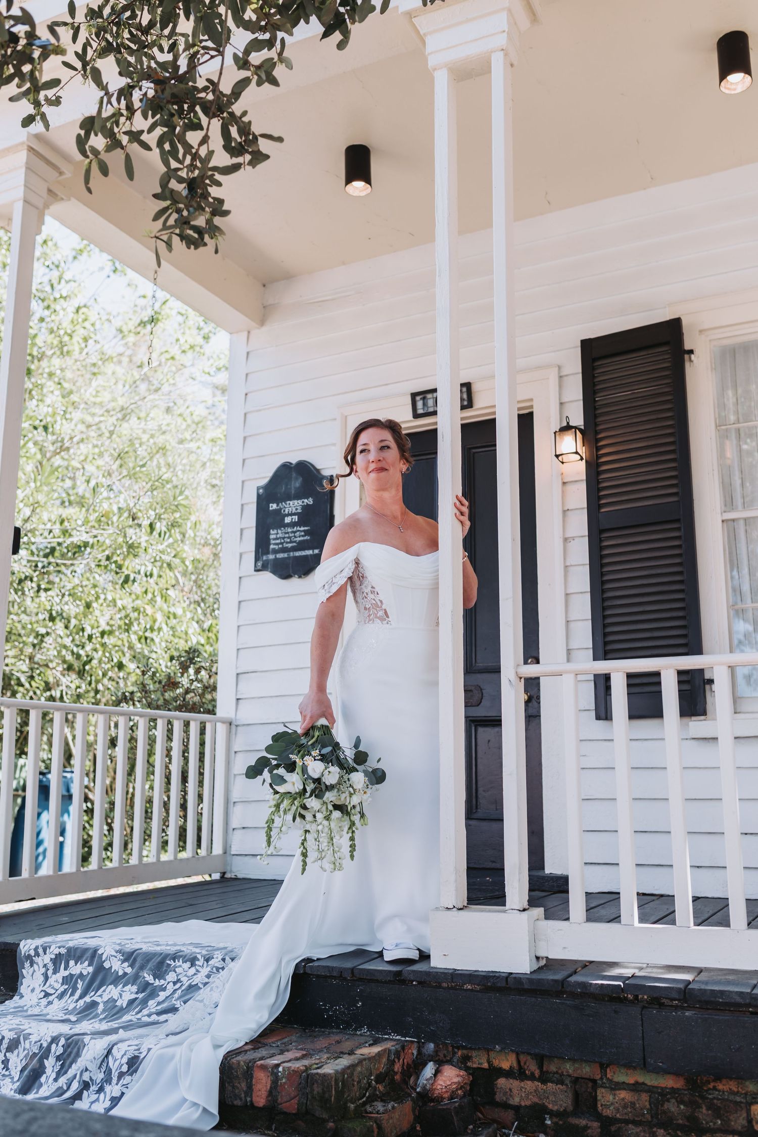 Bride in off-shoulder white gown holding bouquet on porch of white cottage with black shutters.