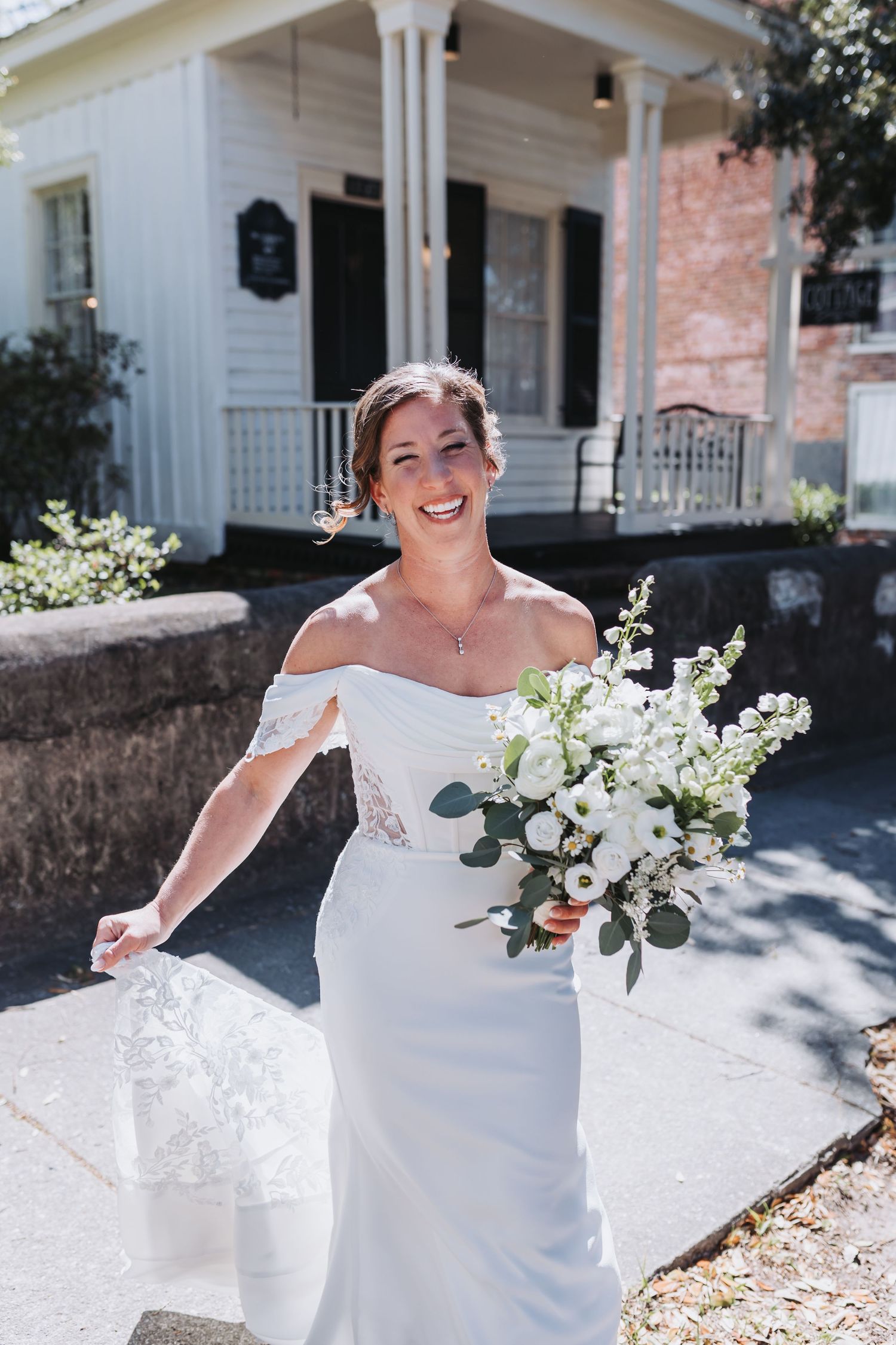 Smiling bride in off-shoulder lace wedding gown carries white floral bouquet outside a charming white house.