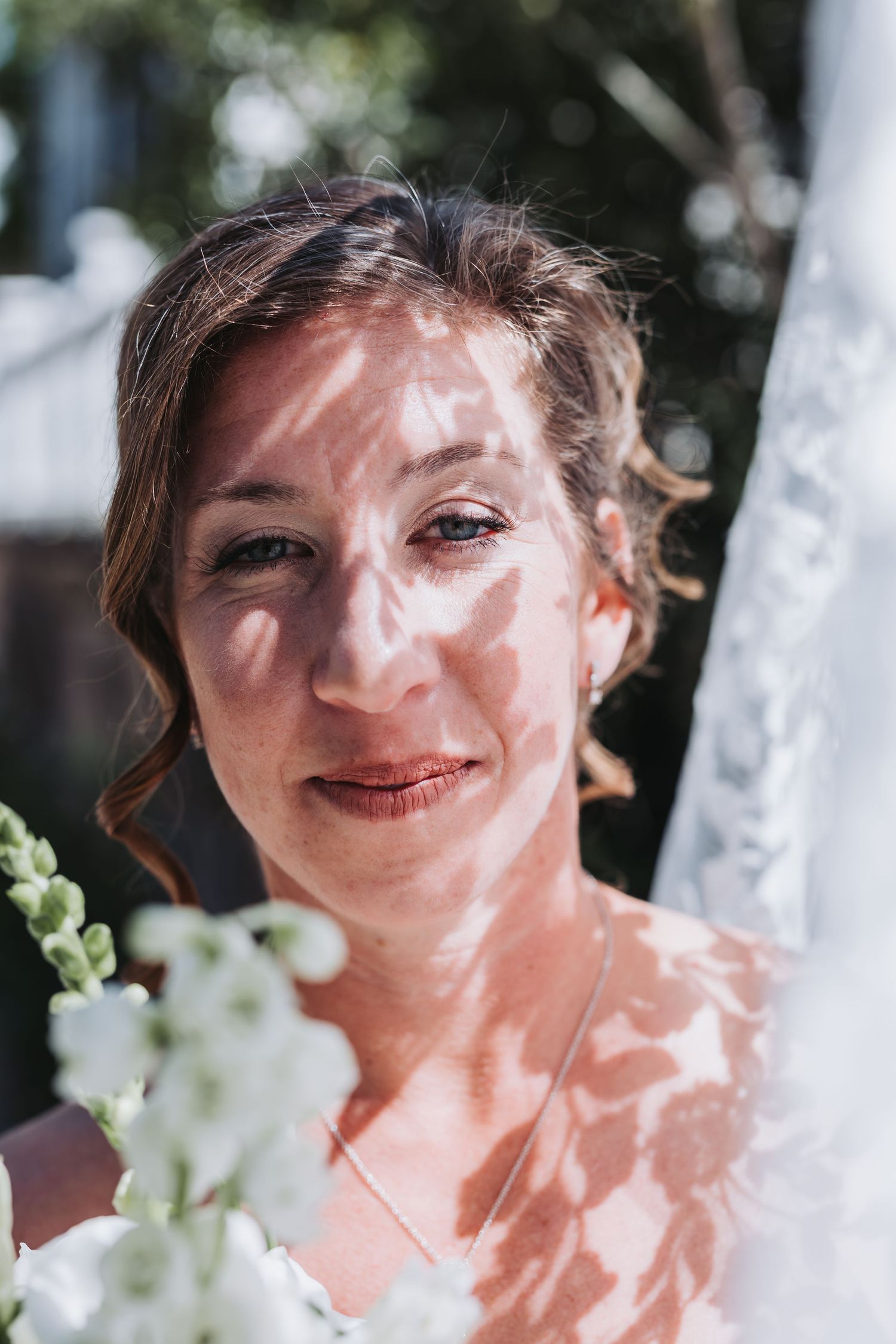 Bride with dappled sunlight on her face holding white flowers outdoors on her wedding day.