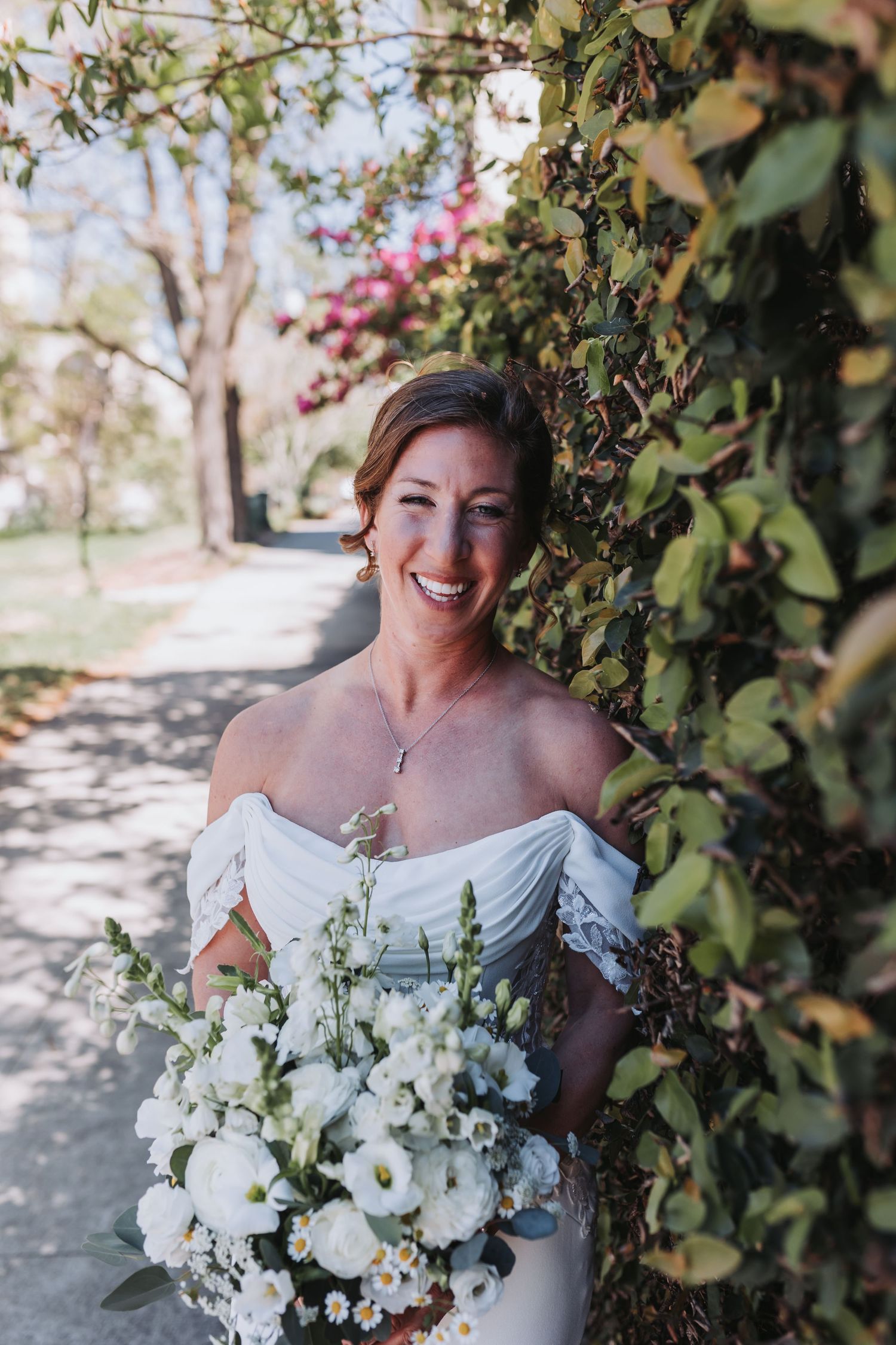 Smiling bride in off-shoulder white gown holds white floral bouquet beside lush ivy wall with pink flowers.