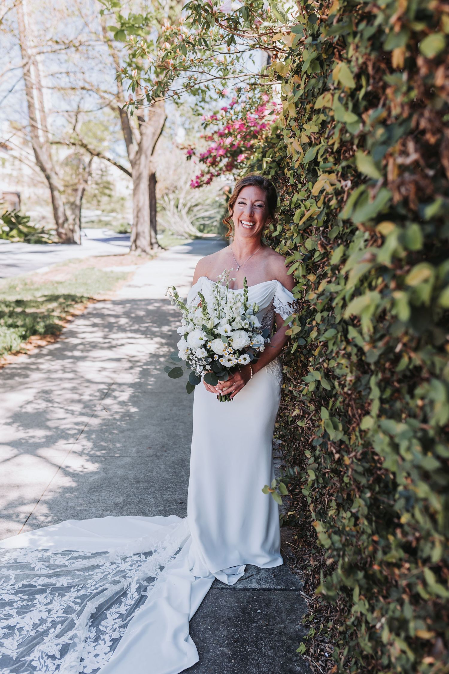 Radiant bride in off-shoulder white gown holds white bouquet beside ivy-covered wall on sunny day.