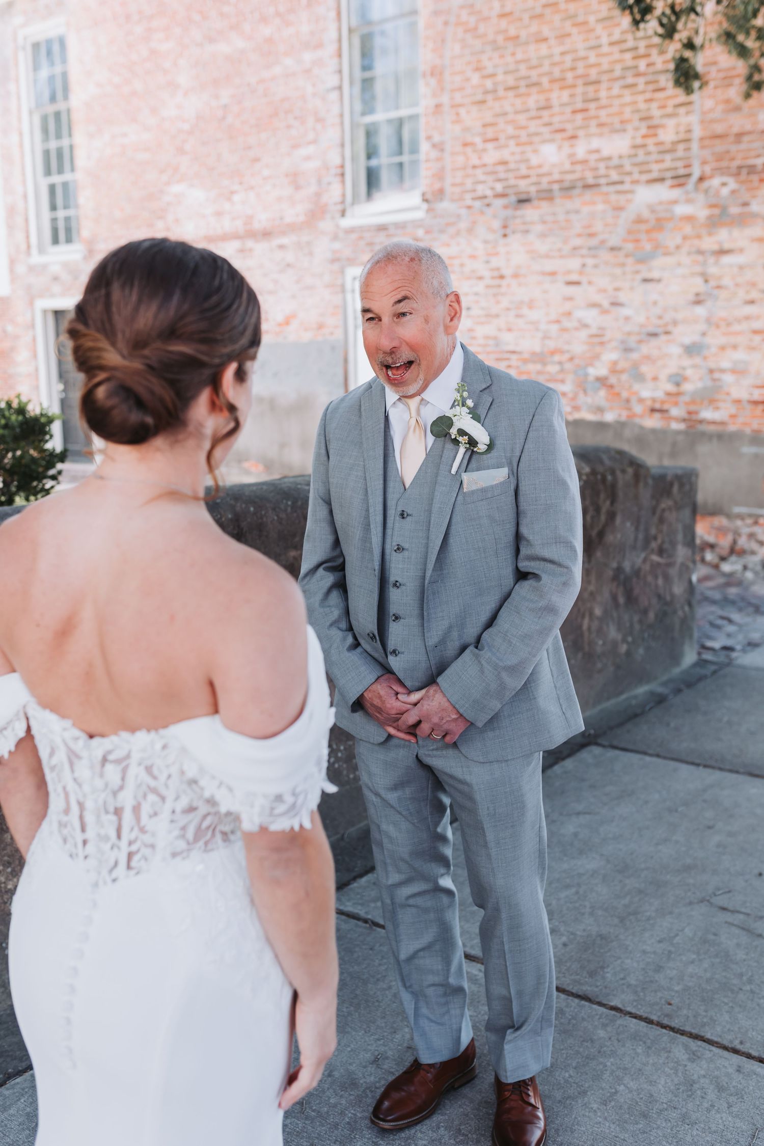 Groom in gray suit reacts with joy seeing bride in white lace gown during first look at brick building.