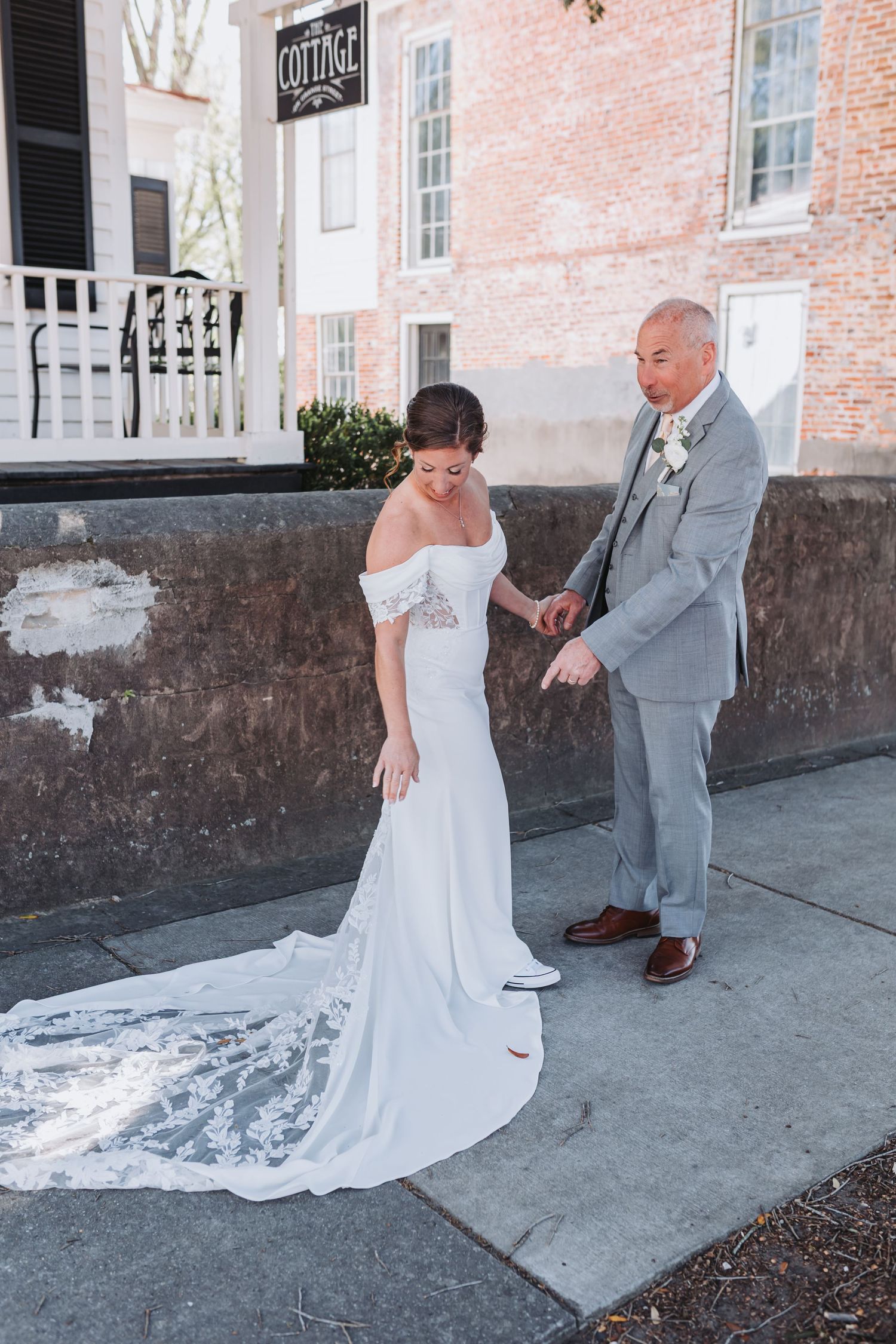 Groom in gray suit adjusting bride's lace train on sidewalk outside The Cottage venue.
