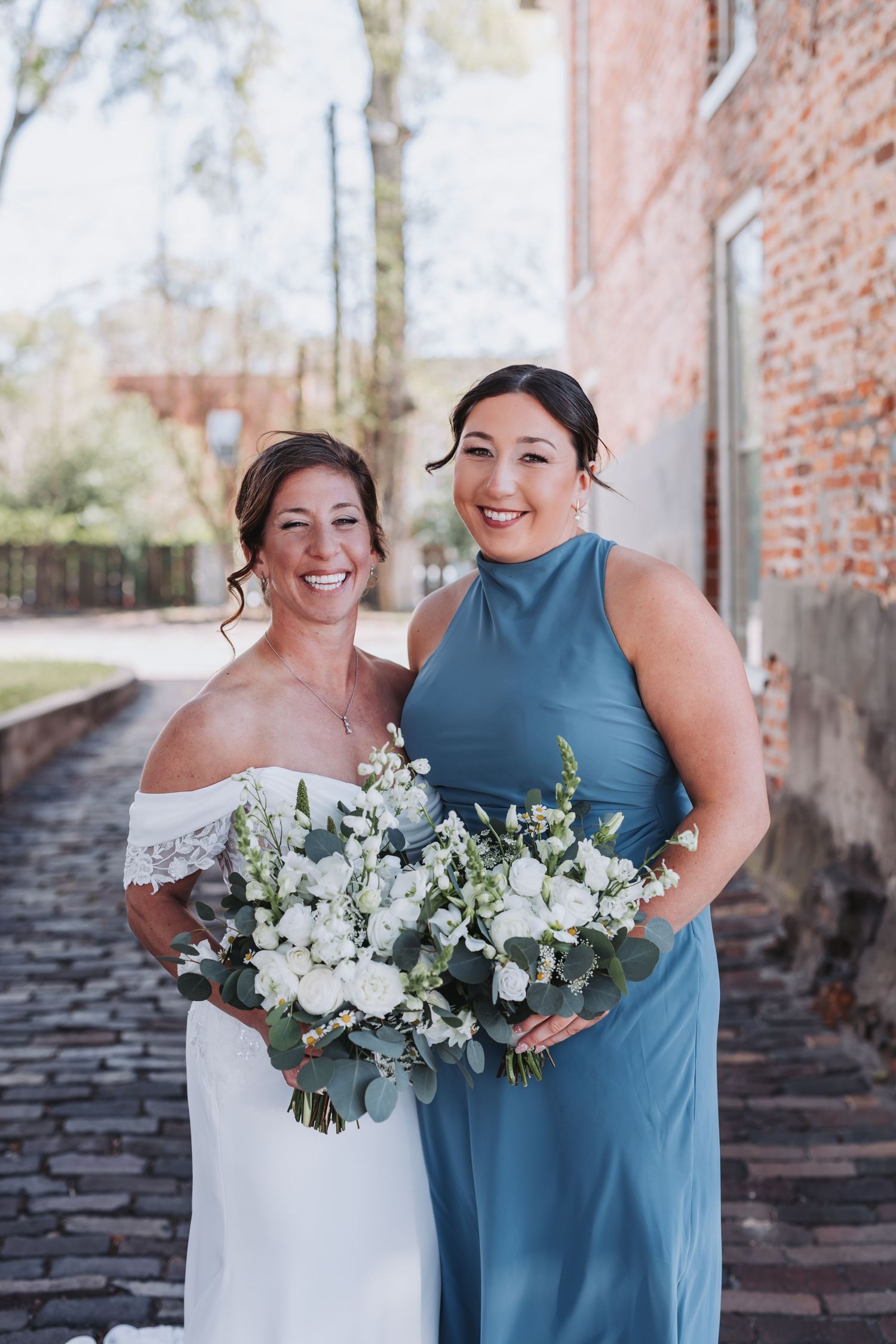 Bride in white off-shoulder gown poses with bridesmaid in blue dress, both holding white bouquets outdoors.