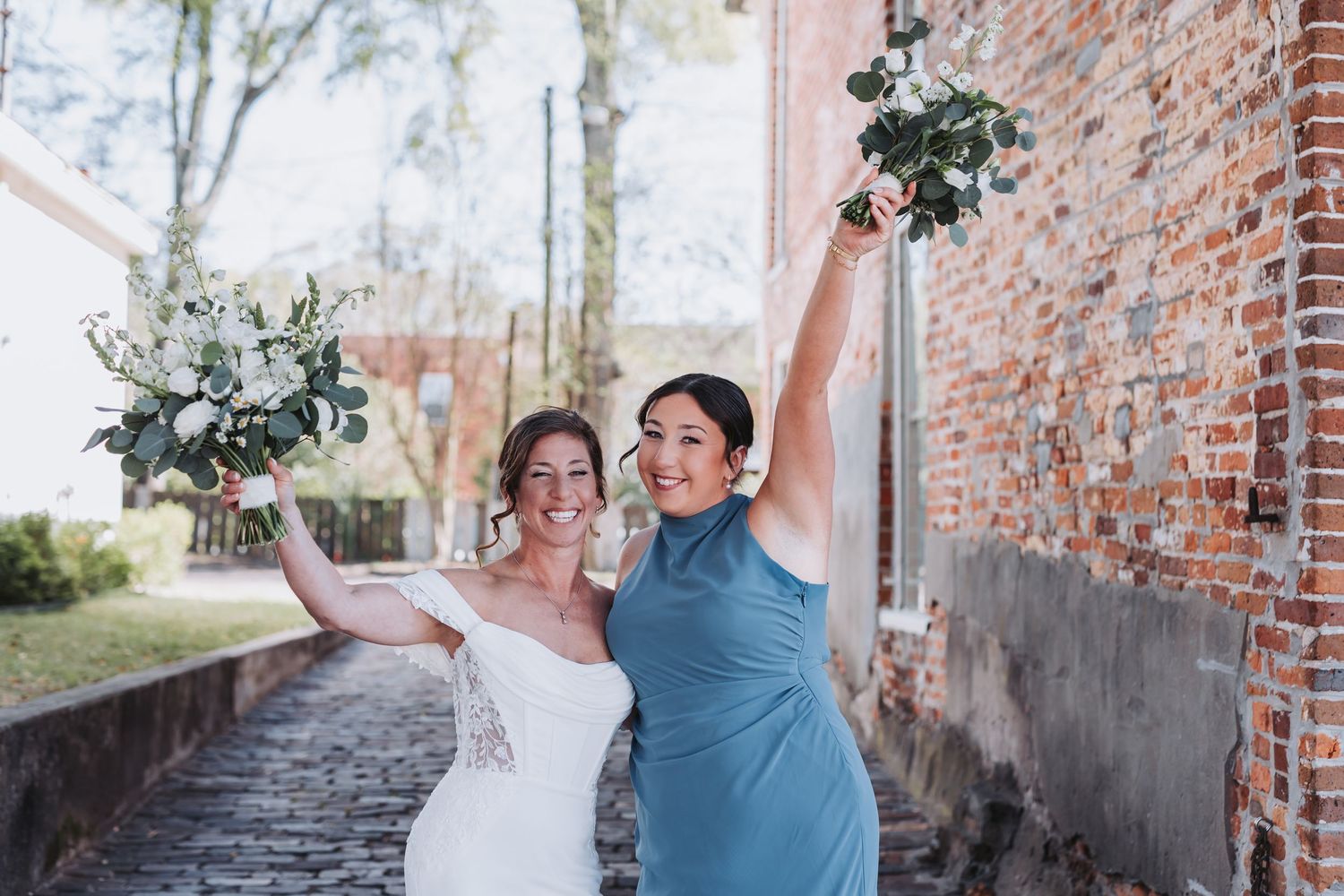 Bride and bridesmaid in blue dress joyfully raising white bouquets near brick wall.