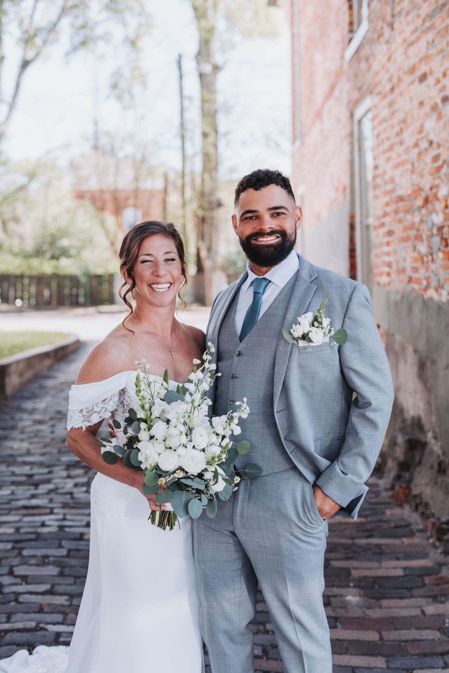 Happy bride and groom pose together, she in white off-shoulder gown, he in light gray suit, near brick wall.