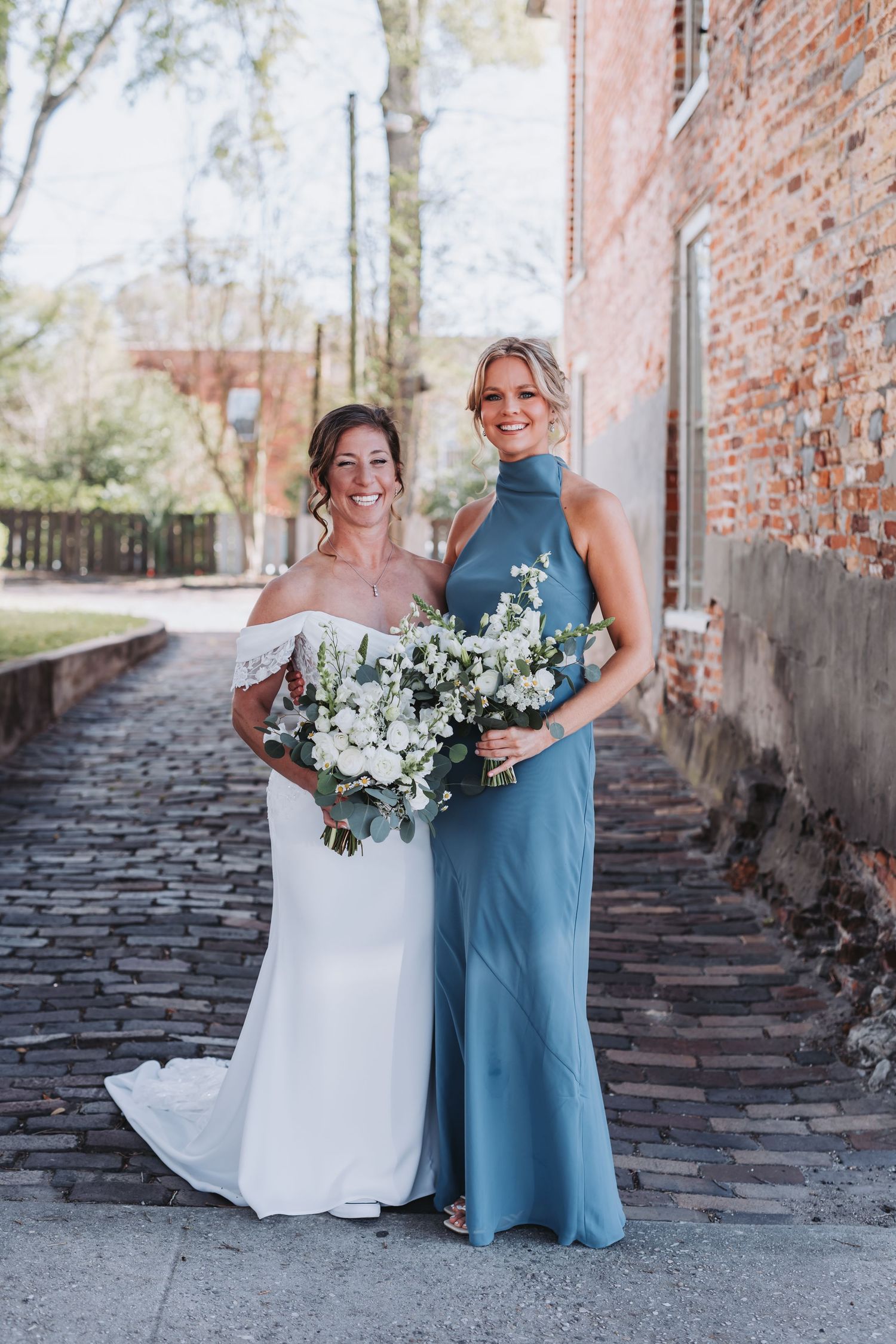 Bride in white off-shoulder gown and bridesmaid in blue halter dress holding bouquets on cobblestone street.