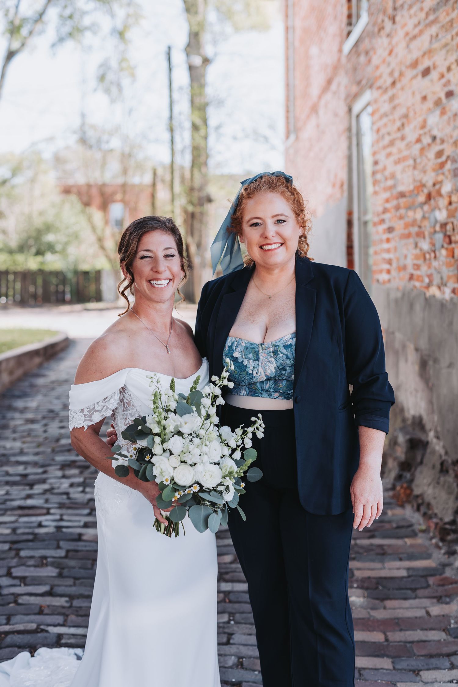 Bride in off-shoulder white gown smiles with red-haired guest in navy blazer on cobblestone path.