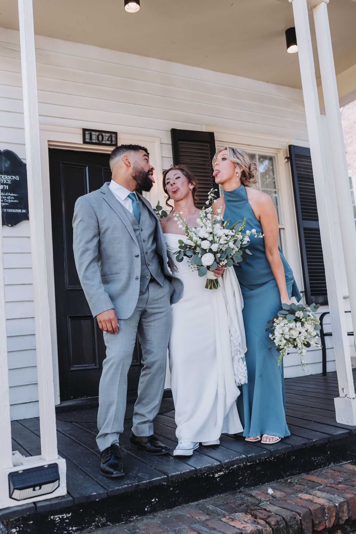 Bride, groom in gray suit, and bridesmaid in blue dress posing on porch of historic building.