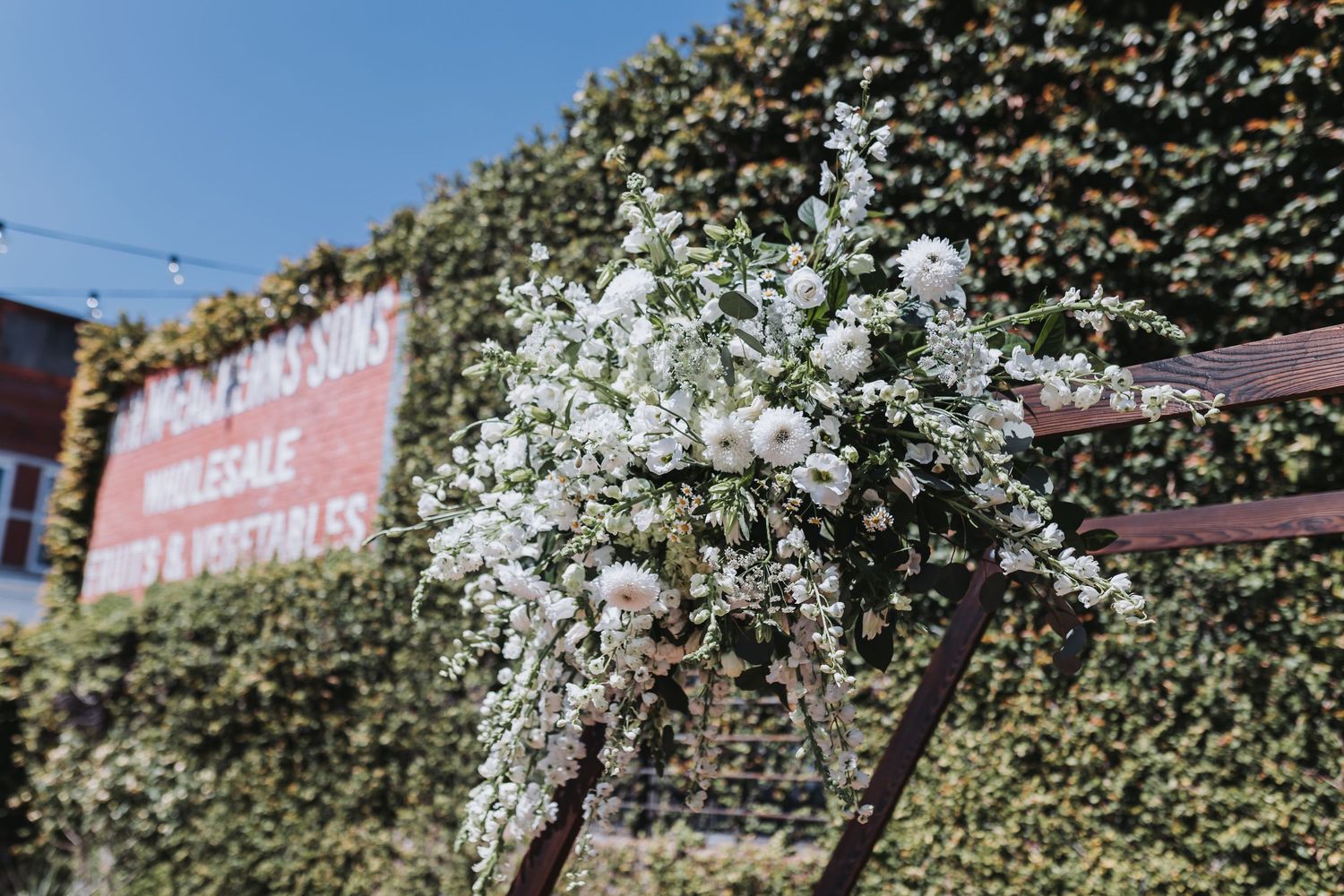 White floral arrangement with roses and baby's breath on a wooden arch against a lush green ivy wall backdrop.
