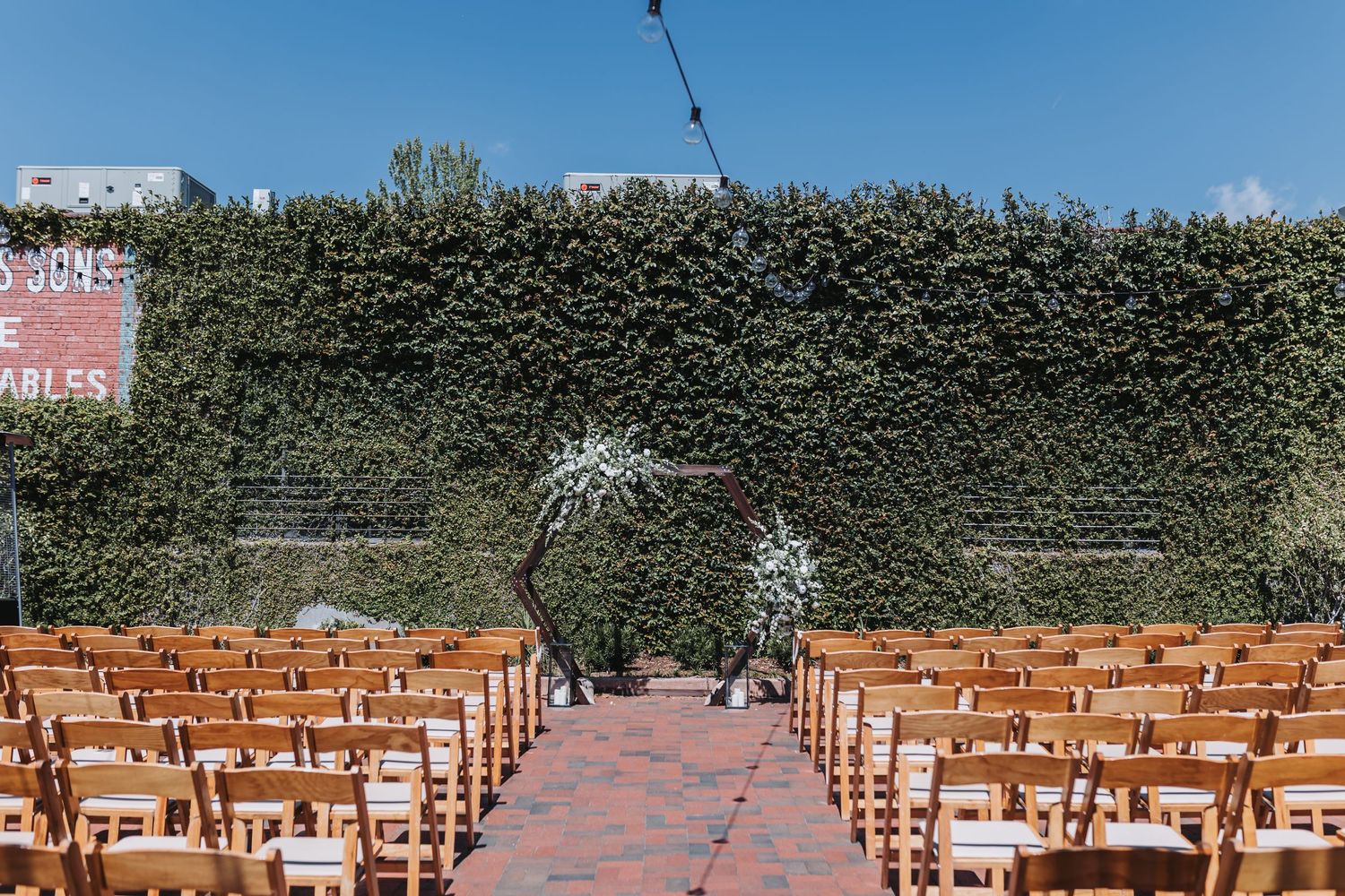 Outdoor wedding ceremony setup with wooden chairs, brick aisle, floral arch, and lush green ivy wall backdrop under blue sky.