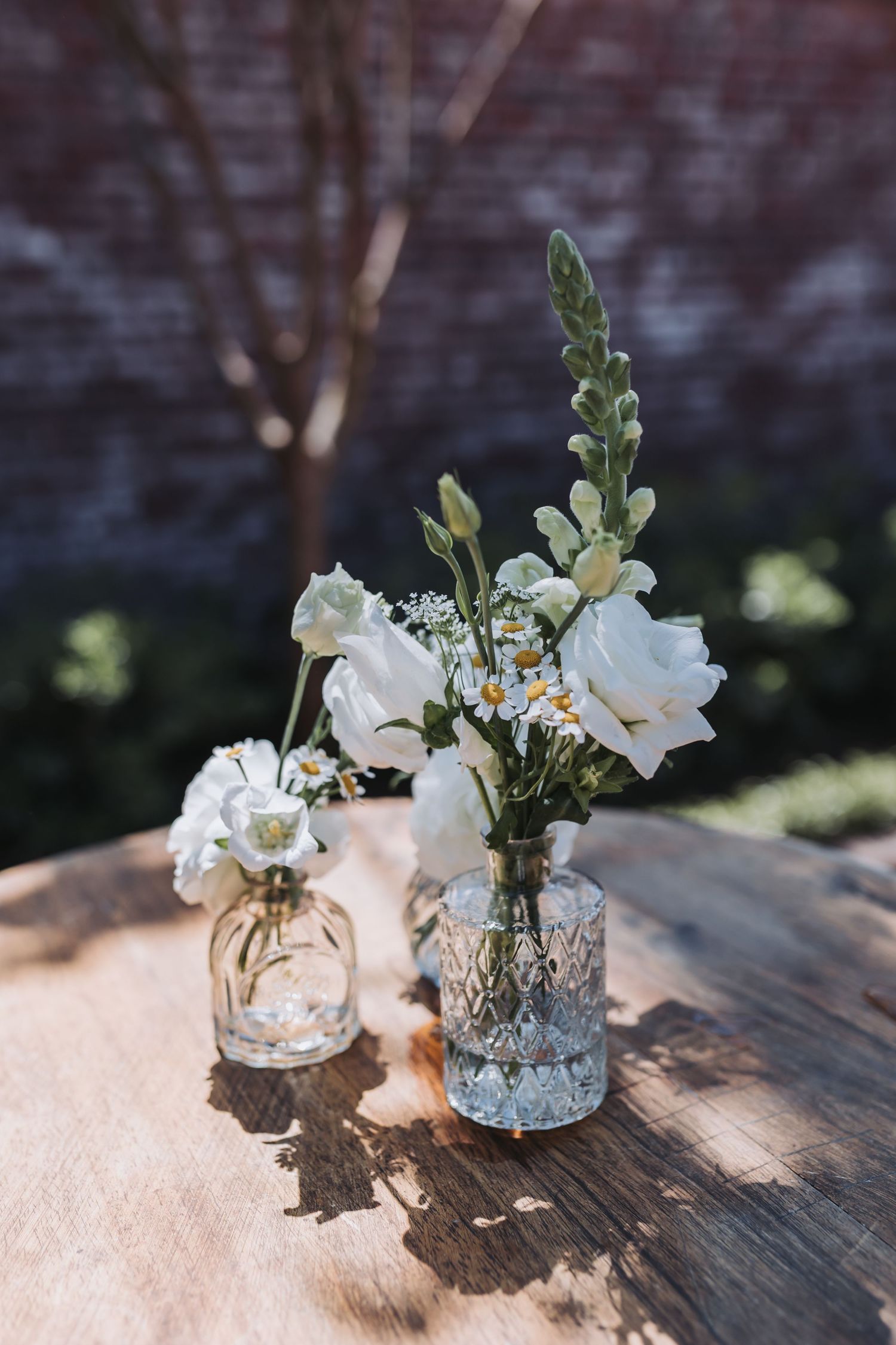 Two glass vases with white flowers and daisies on a rustic wooden table outdoors in sunlight.