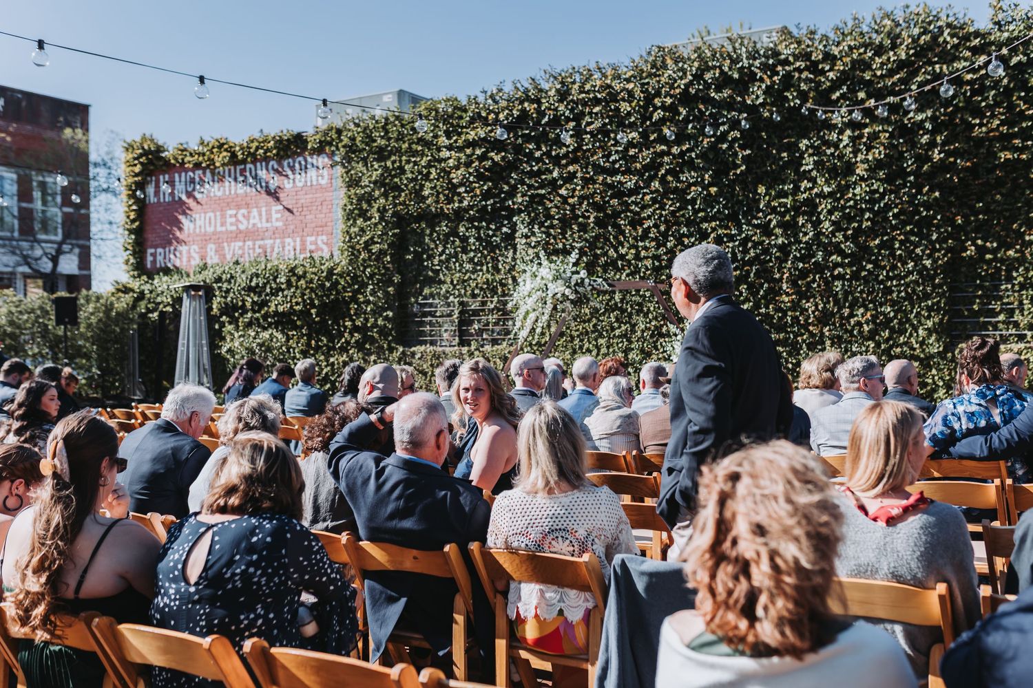 Outdoor wedding ceremony with guests seated in wooden chairs facing a speaker, surrounded by lush green ivy walls.