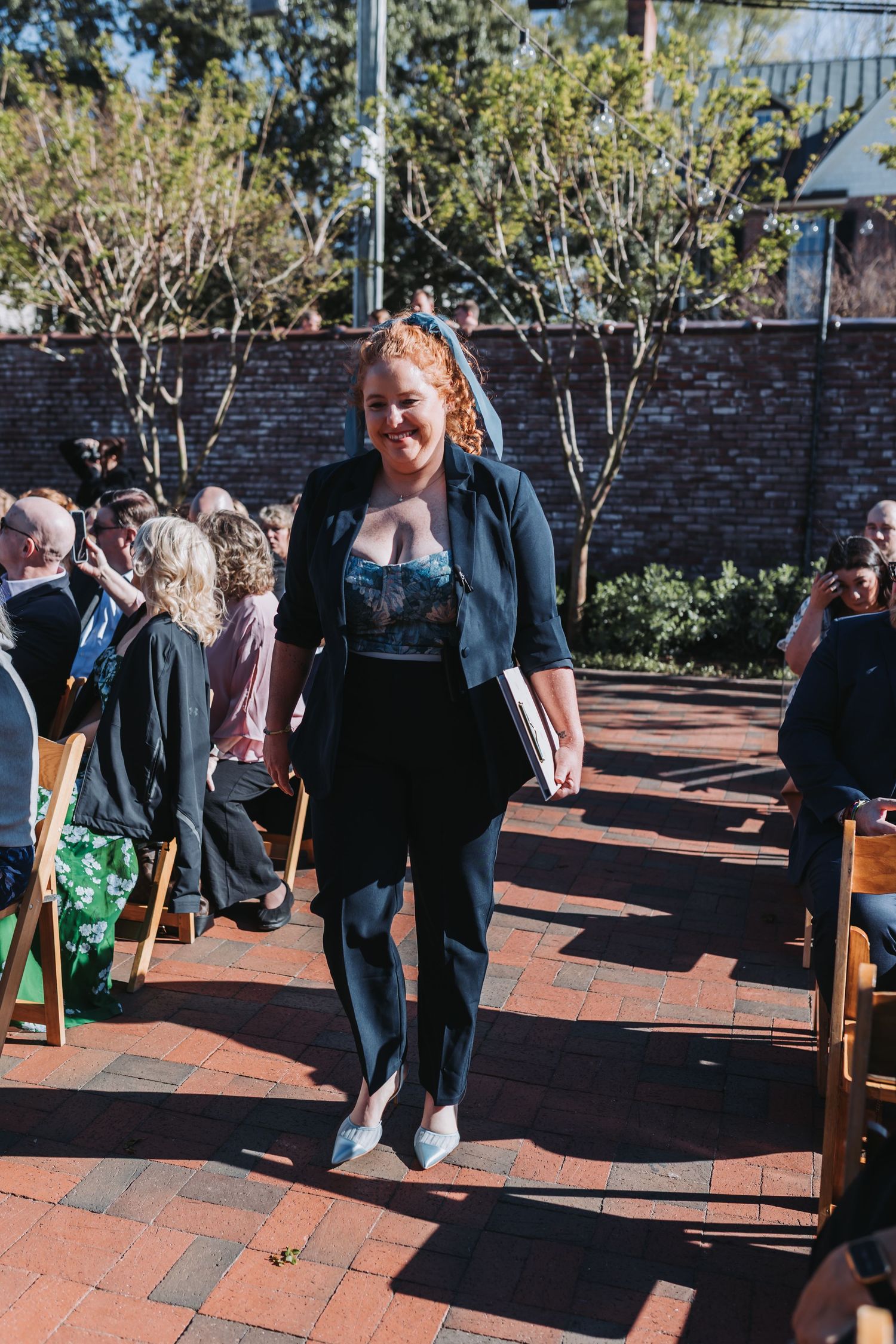 Woman in black blazer and lace top walks down outdoor wedding aisle on brick pathway.