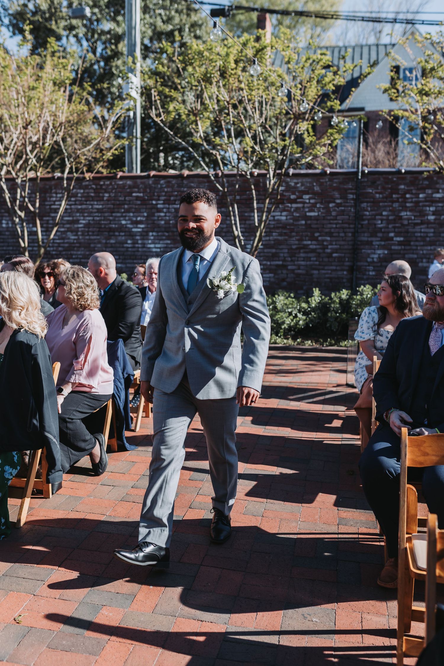 Groom in gray suit with boutonniere walks down brick aisle at outdoor wedding ceremony.