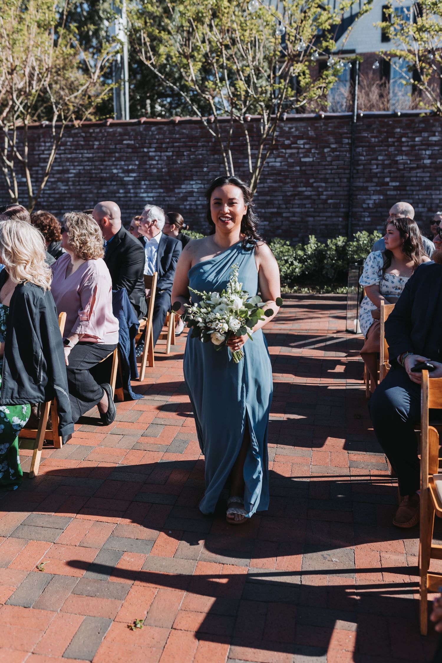 Smiling bridesmaid in teal gown walks down brick aisle holding white bouquet at outdoor wedding ceremony.