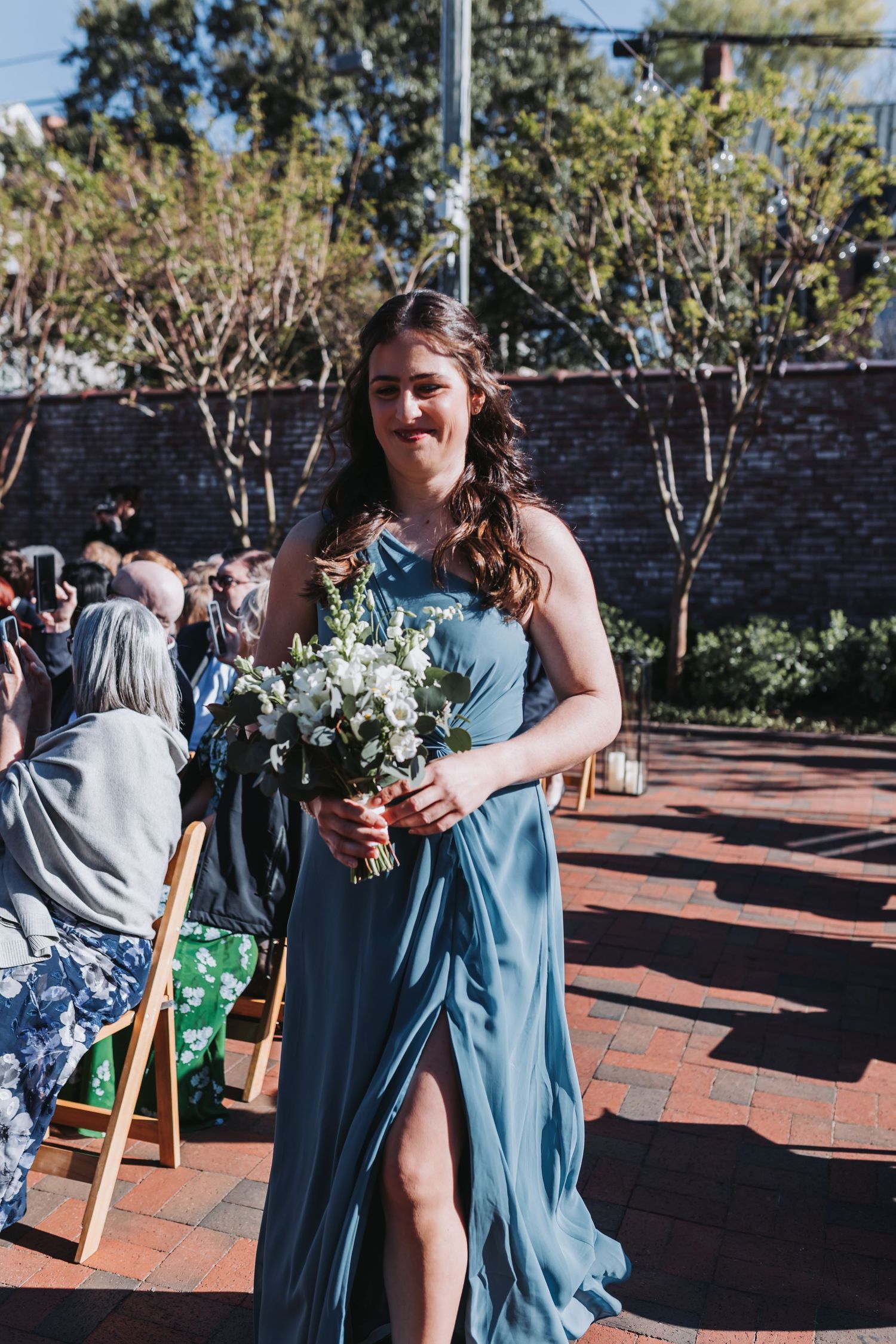 Bridesmaid in blue dress holds white floral bouquet walking down outdoor wedding aisle.