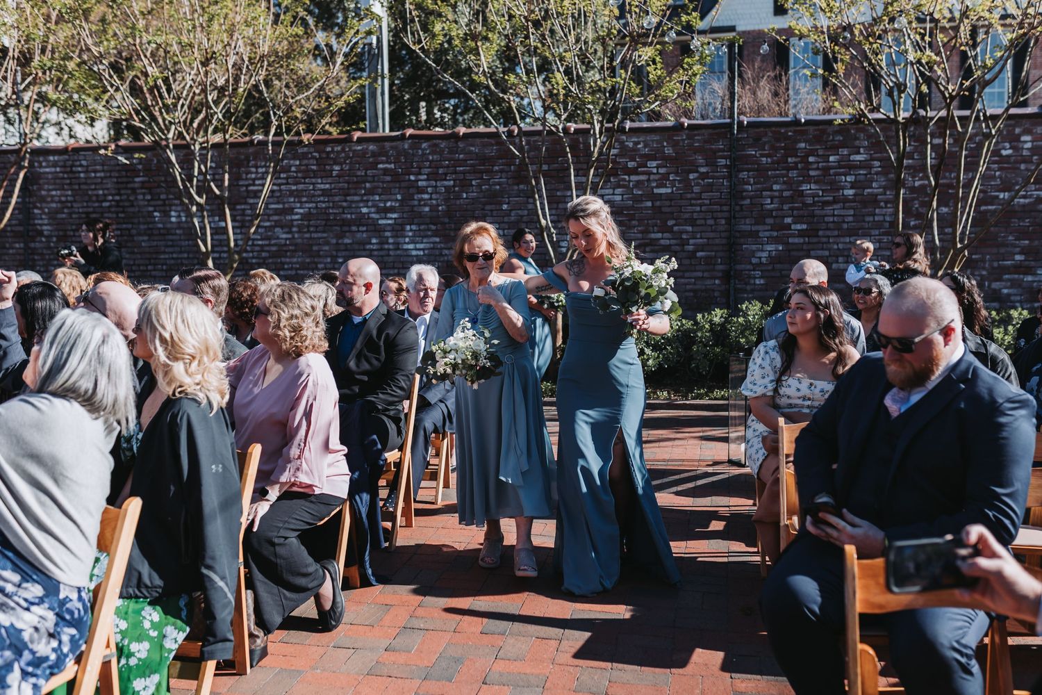 Bridesmaids in blue dresses walk down brick aisle at outdoor wedding ceremony with seated guests.