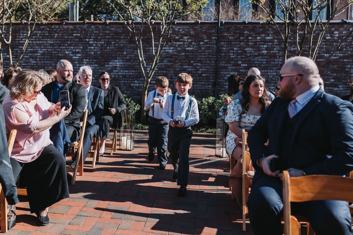 Young ring bearer walks down brick outdoor wedding aisle as seated guests smile and applaud during sunny ceremony.