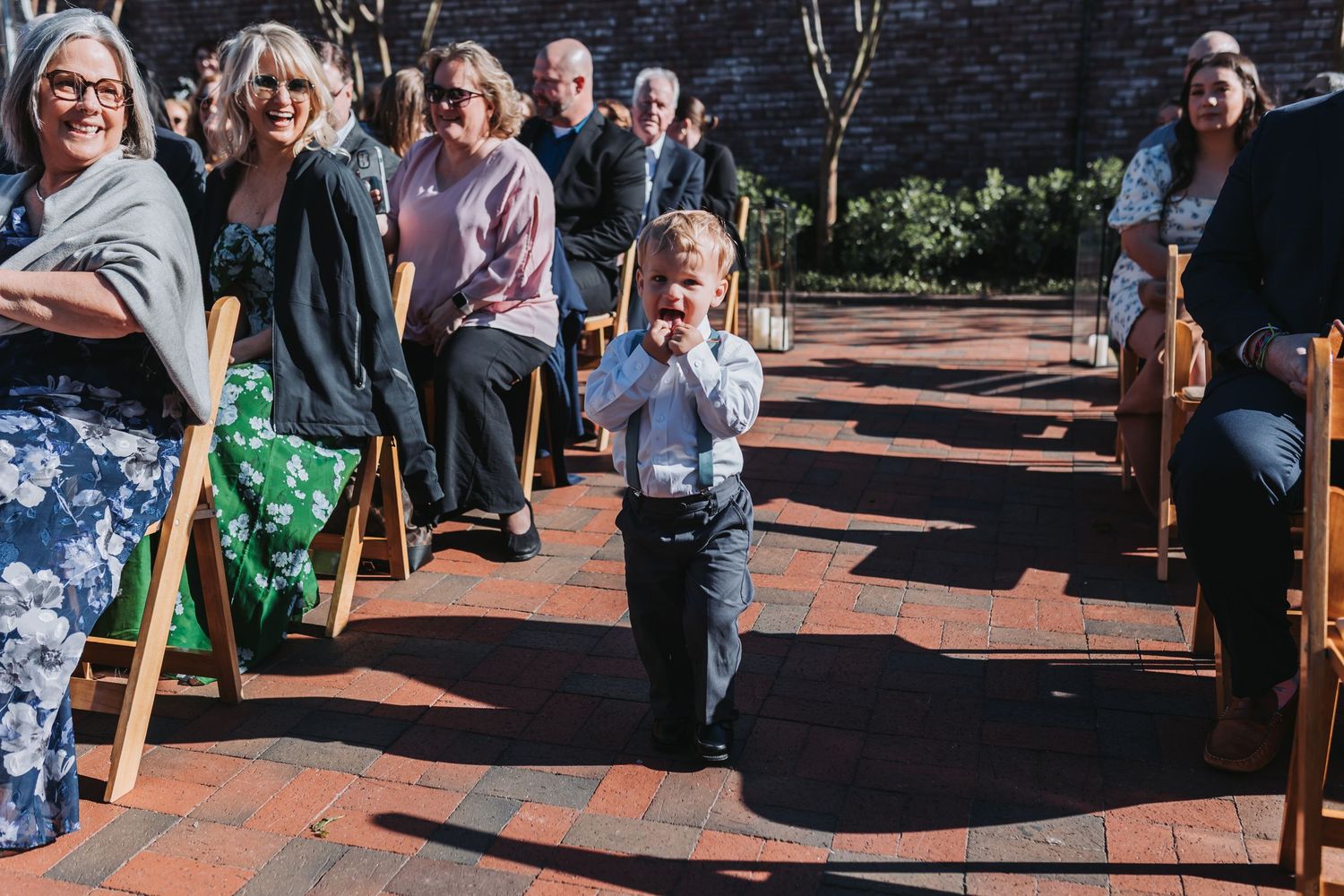 Adorable ring bearer walks down outdoor wedding aisle amid smiling seated guests on brick patio.