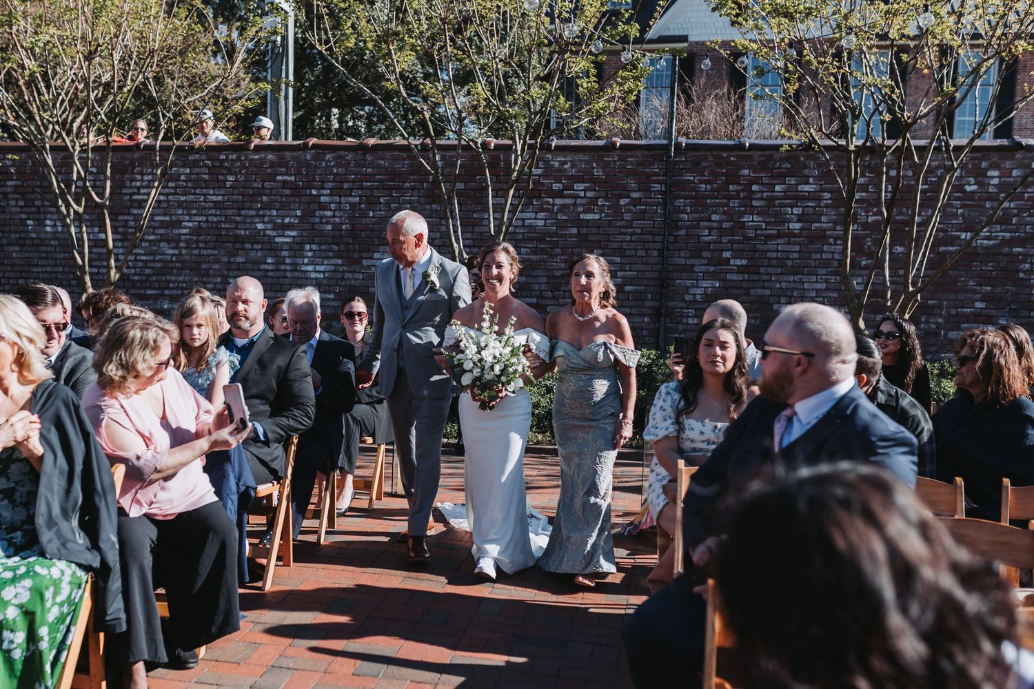 Bride walks down brick aisle with escort at outdoor wedding ceremony surrounded by seated guests.