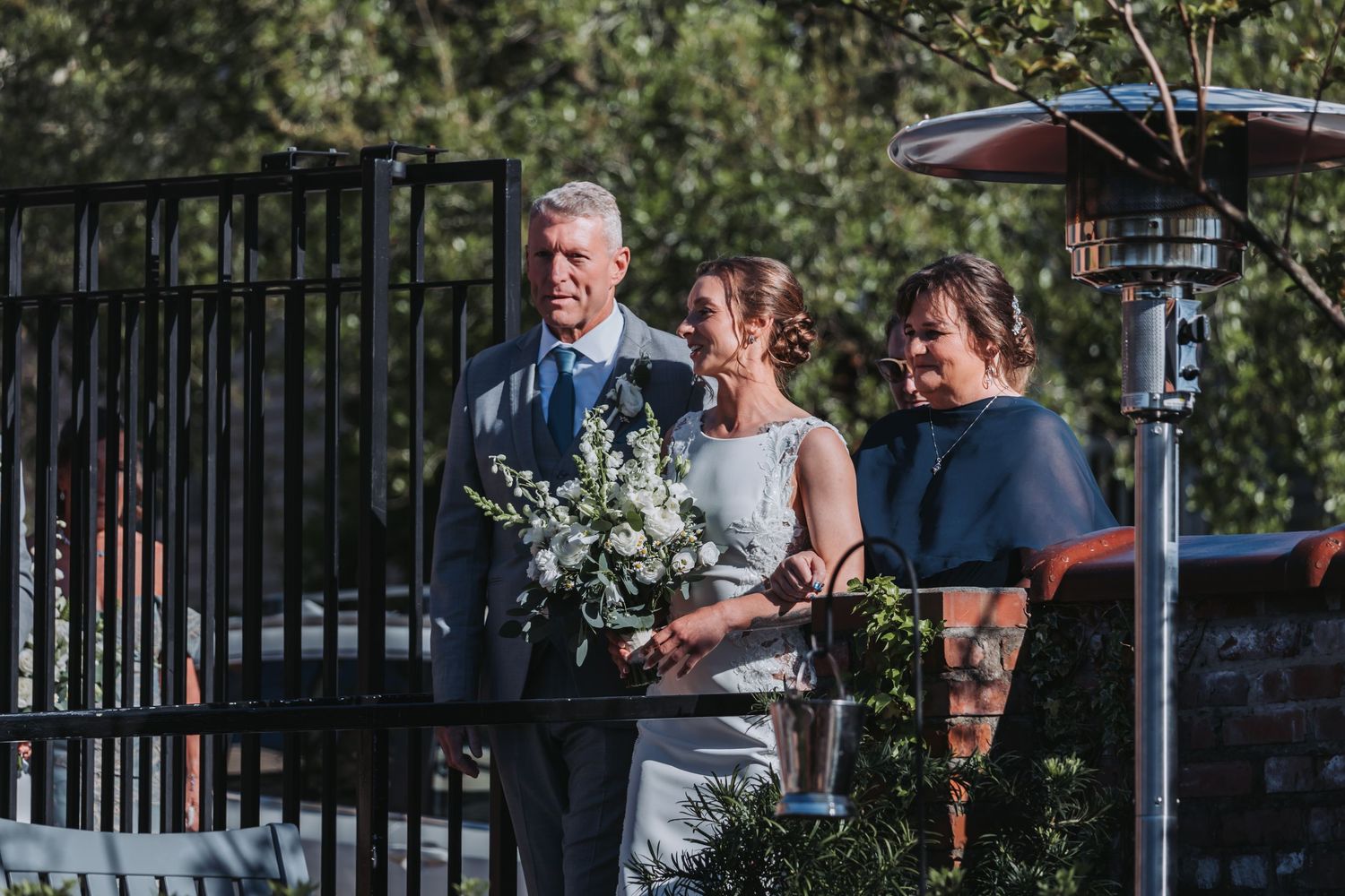 Bride holding white bouquet stands with two guests at outdoor wedding venue with iron gate and trees.