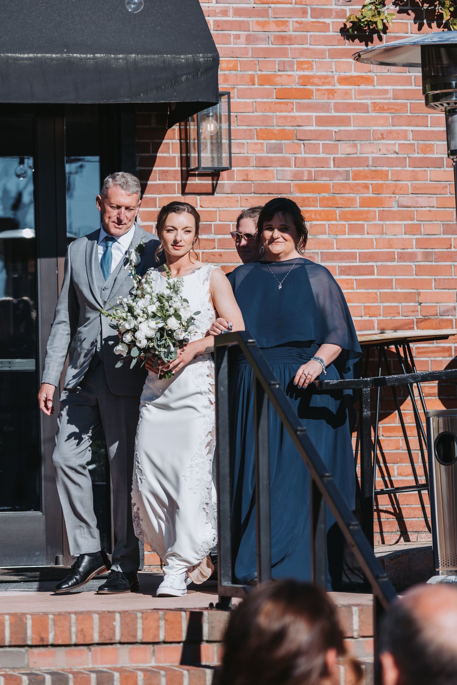 Bride in white gown holding bouquet descends brick steps flanked by father in gray suit and mother in navy dress.