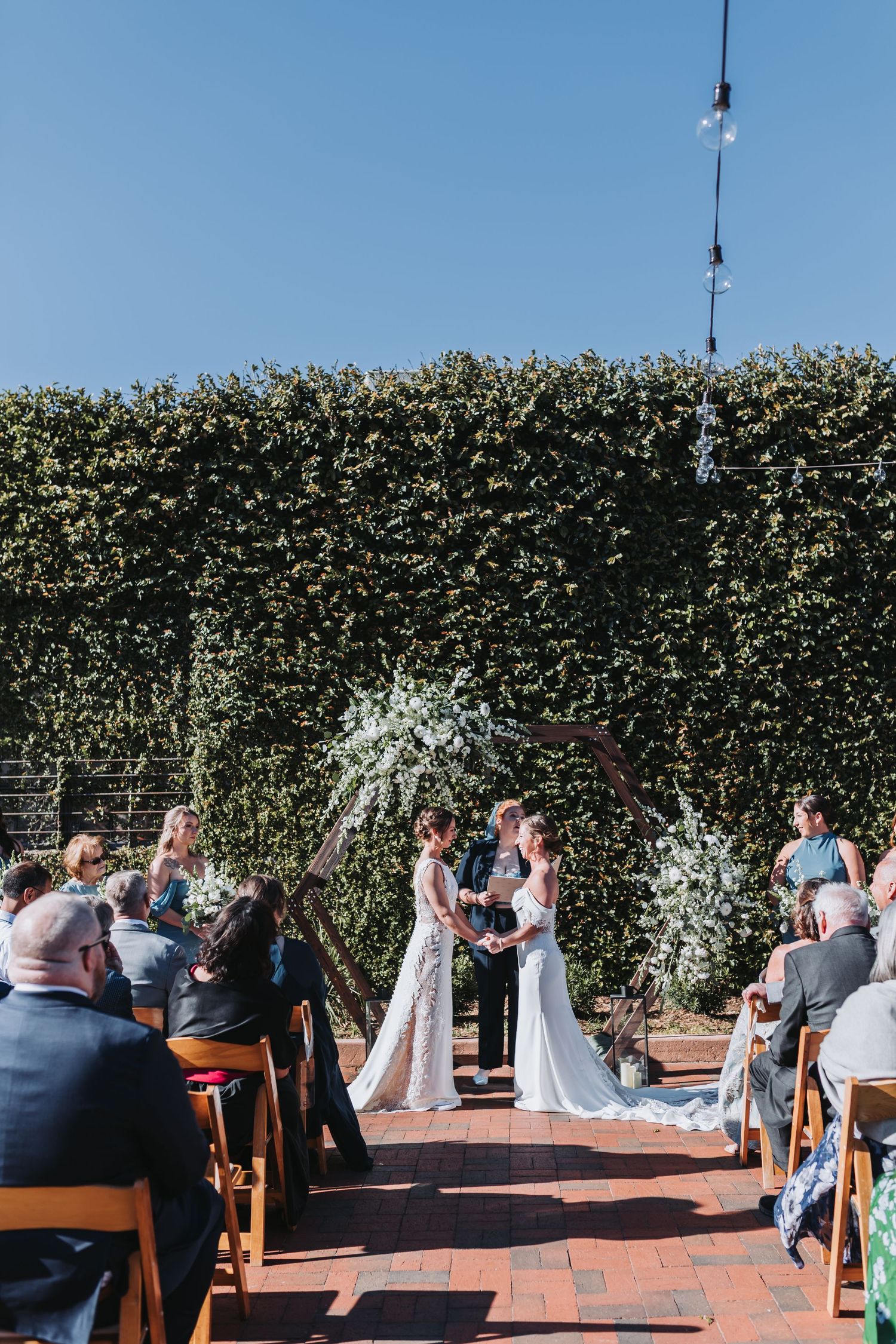 Two brides exchange vows at outdoor wedding ceremony beneath floral arch against lush green ivy wall.