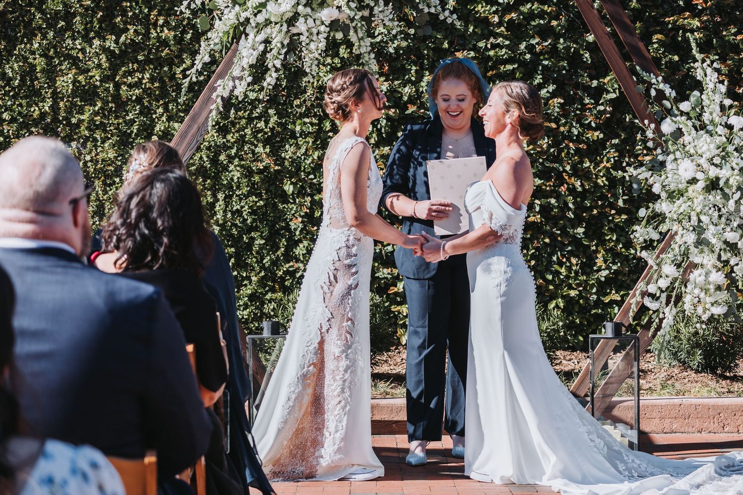 Two brides exchange vows before officiant under floral arch at sunny outdoor wedding ceremony.
