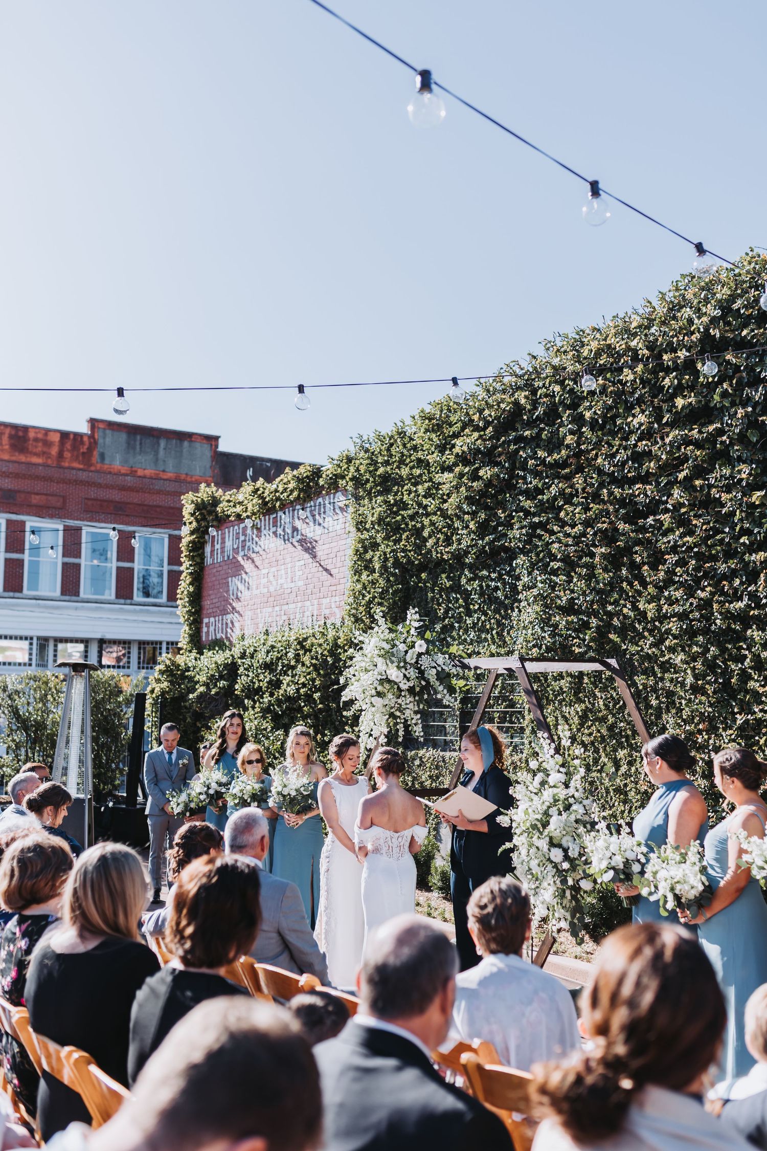 Two brides hold hands at altar during outdoor wedding ceremony with ivy wall, floral arch, and string lights above.