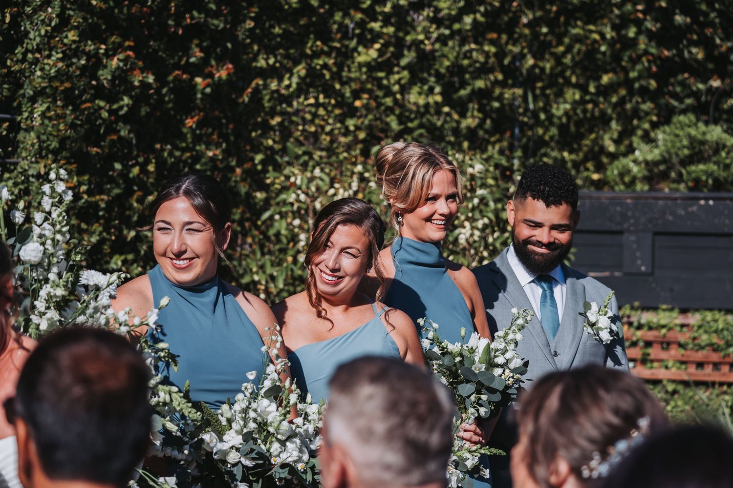 Bridesmaids in dusty blue dresses holding white floral bouquets smile during an outdoor wedding ceremony.