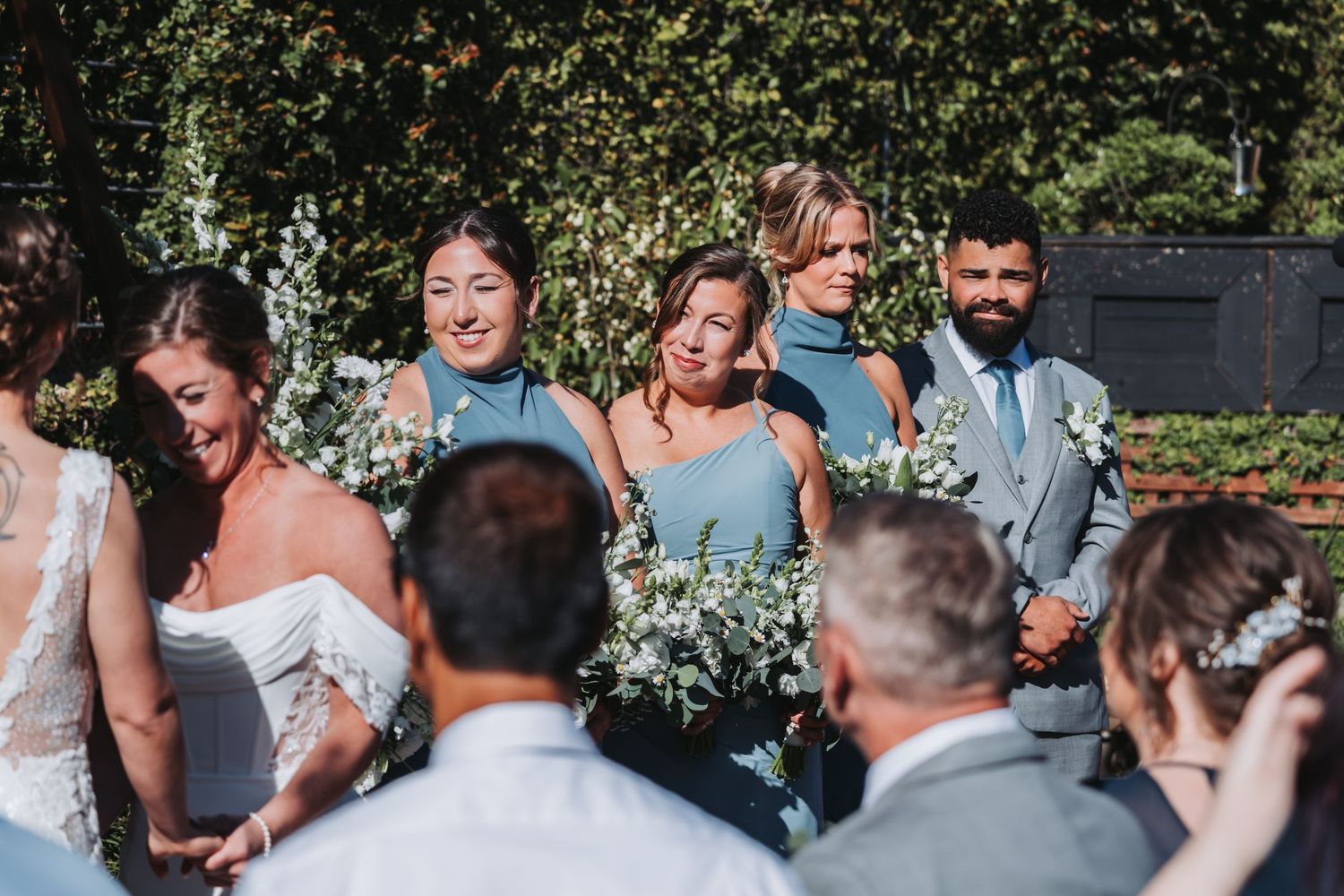 Bridesmaids in dusty blue gowns hold white bouquets beside bride at outdoor wedding ceremony.