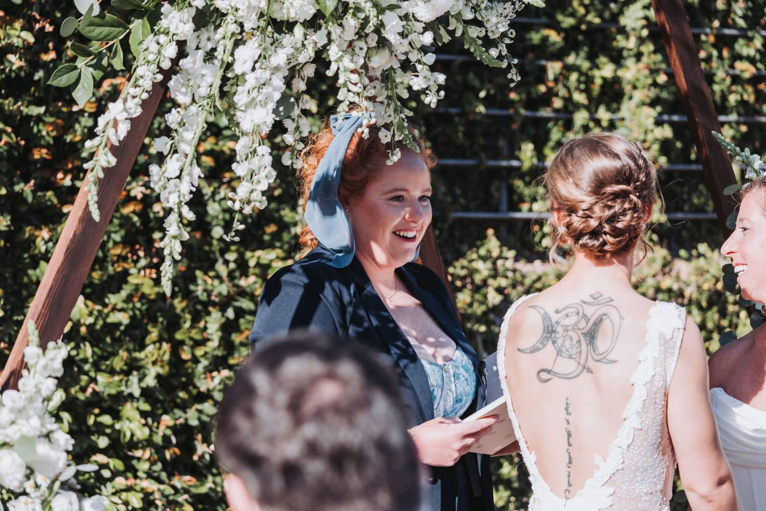 Smiling wedding officiant in blue dress under floral arch with tattooed bride in white lace gown at outdoor ceremony.