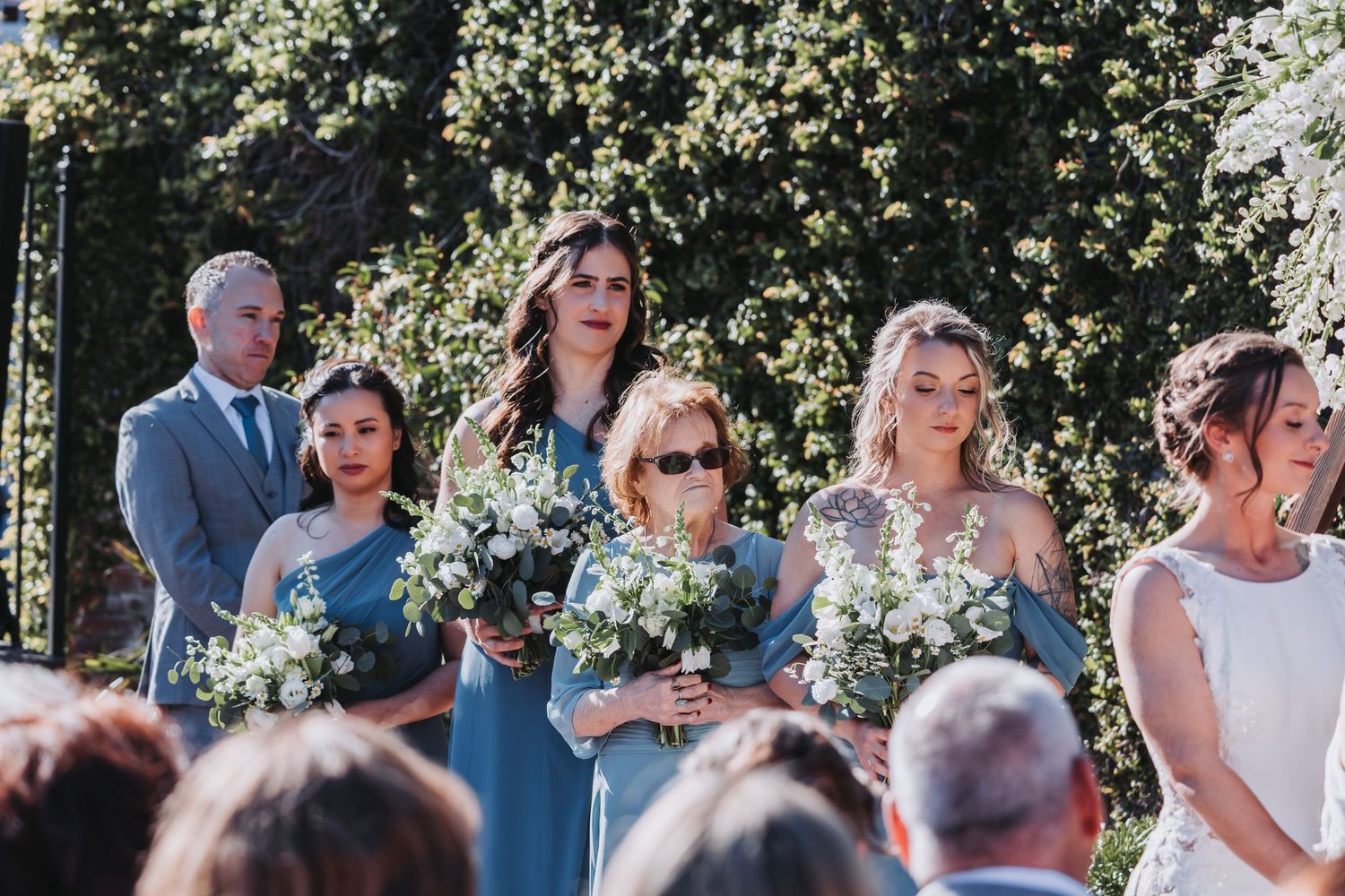 Bridesmaids in dusty blue dresses holding white bouquets stand during outdoor wedding ceremony.