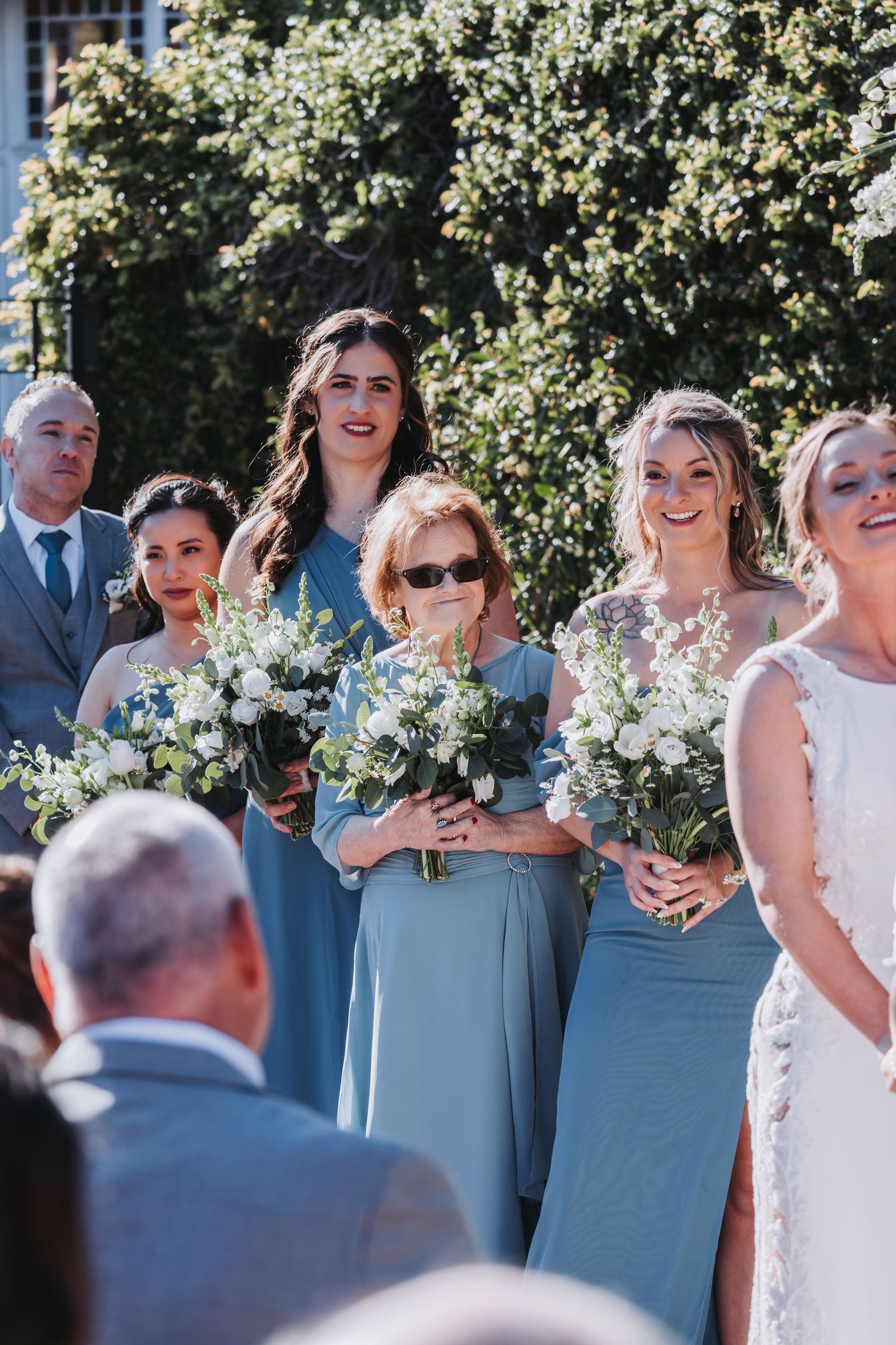Bridesmaids in dusty blue dresses holding white floral bouquets stand with an older woman during an outdoor wedding ceremony.