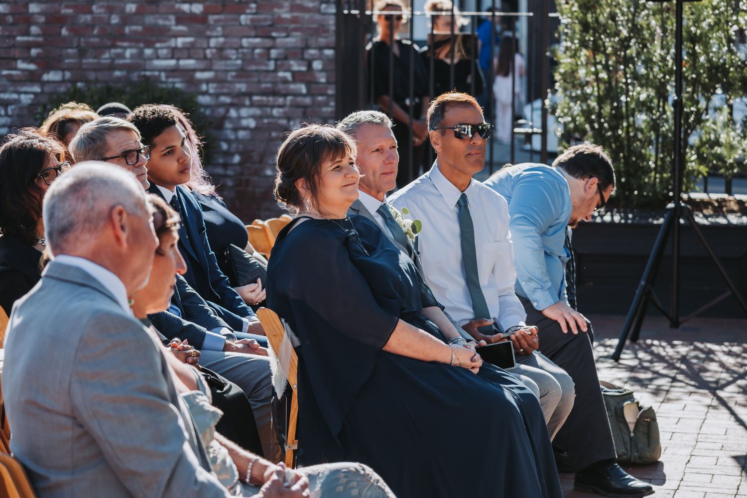 Wedding guests seated on wooden chairs watch outdoor ceremony in sunlight, woman in dark top and man in sunglasses.