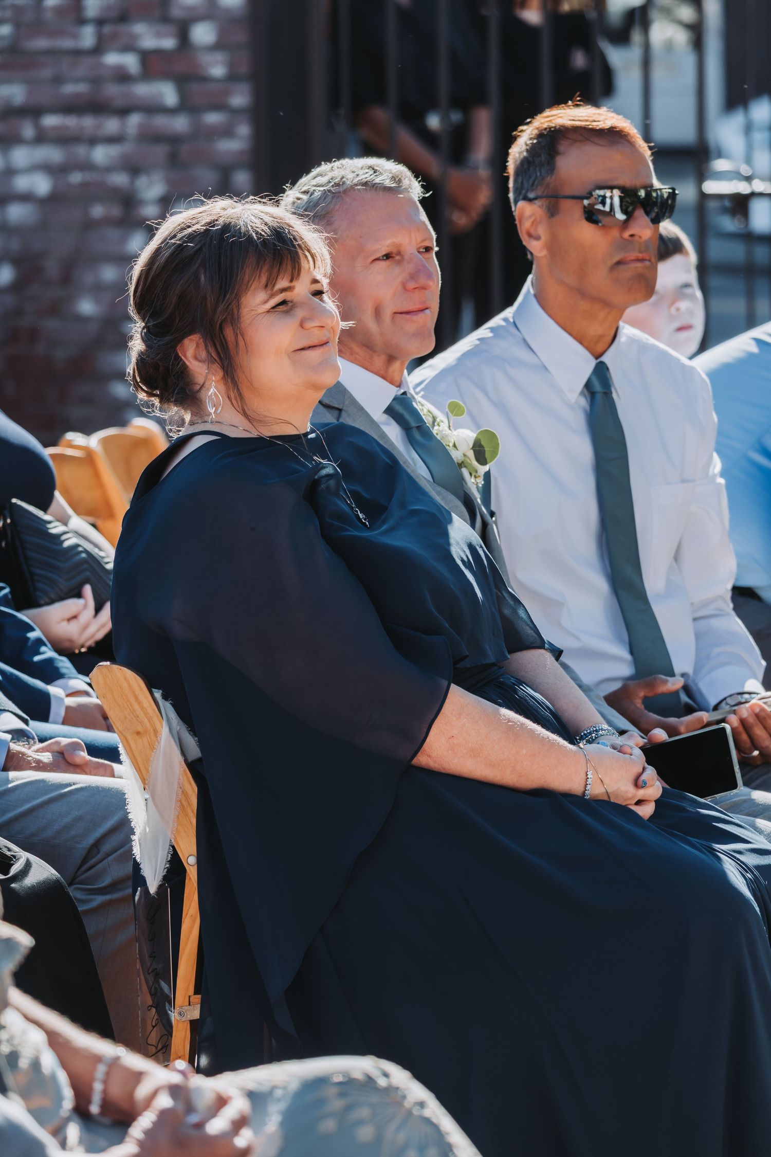 Wedding guests in formal attire seated outdoors watching ceremony on sunny day.