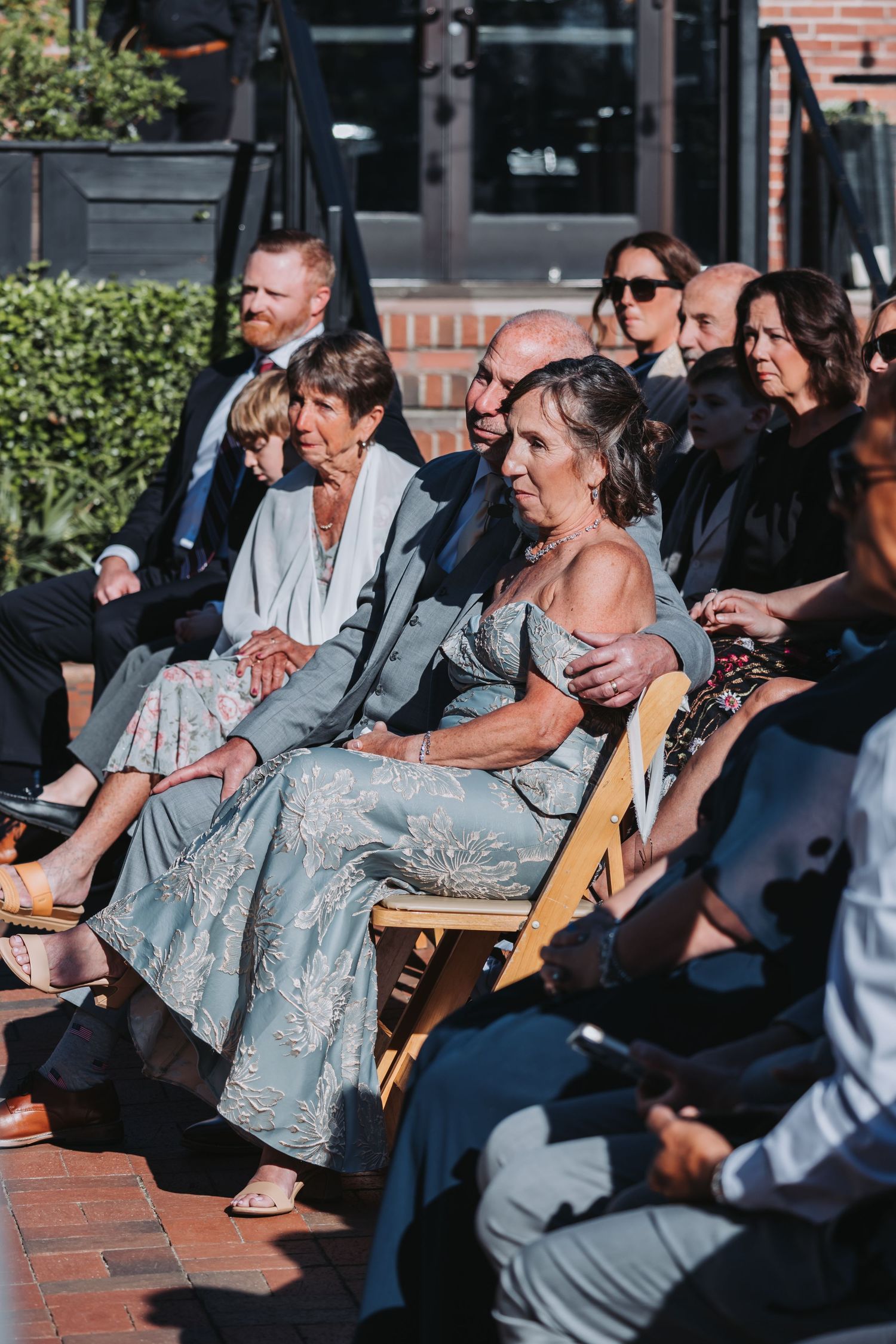 Wedding guests seated outdoors in wooden chairs watching ceremony on sunny day.