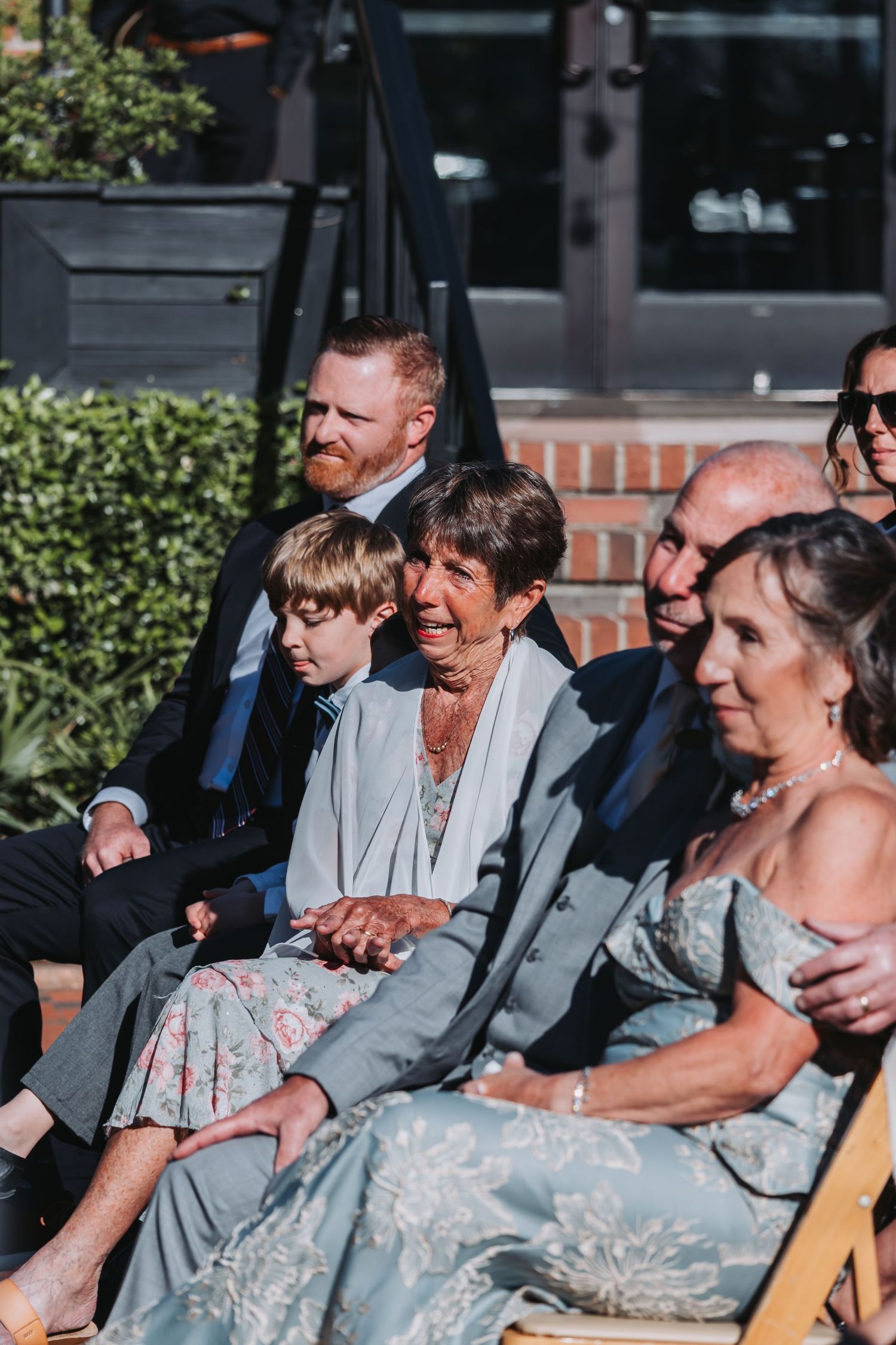 Emotional wedding guests seated outdoors, including an elderly woman and child in blue-gray attire, watching a ceremony.